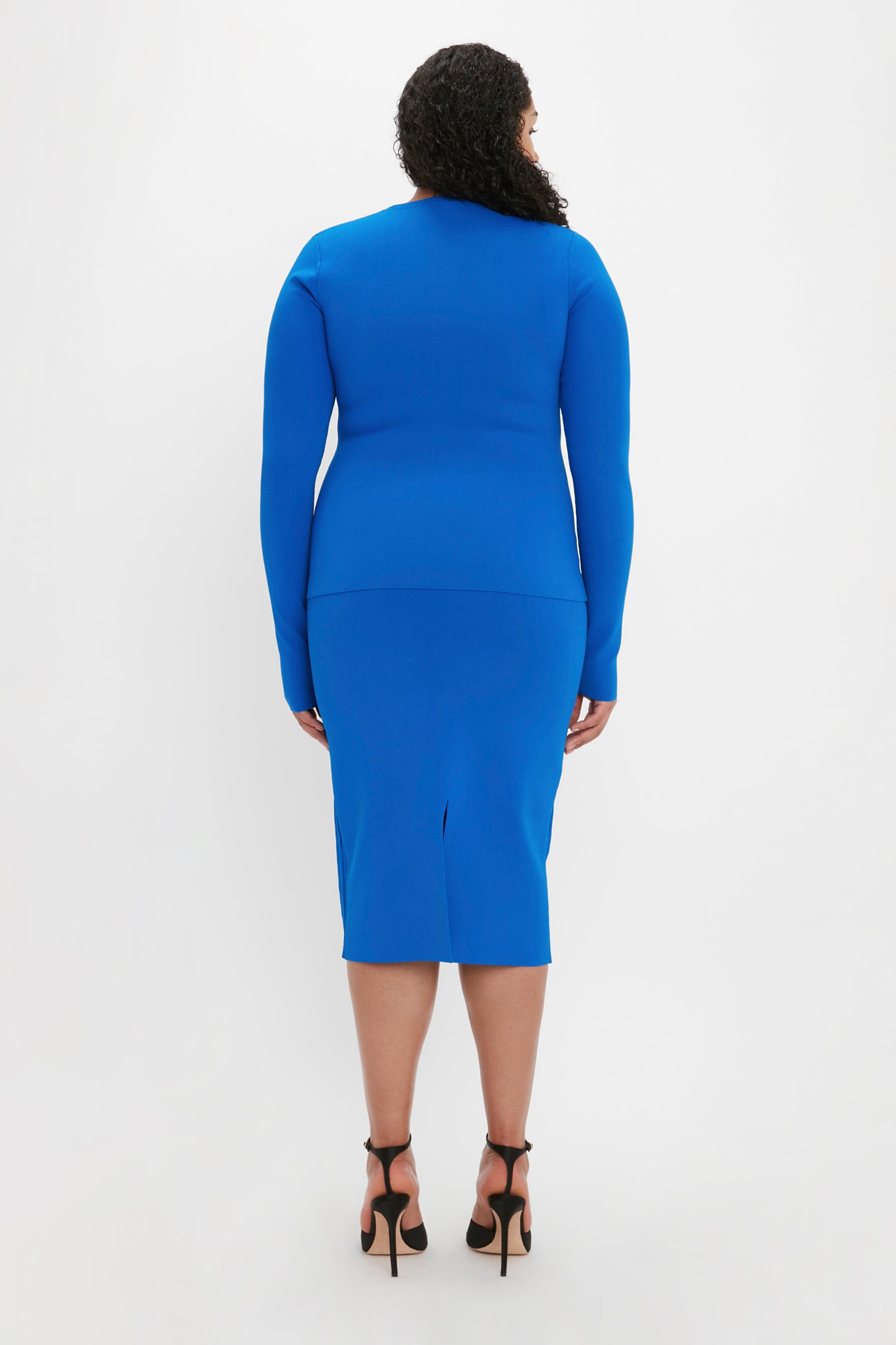 A person with curly hair is seen from the back wearing a form-fitting VB Body Front Slash Crew Neck Top in Cobalt by Victoria Beckham and black high heels.