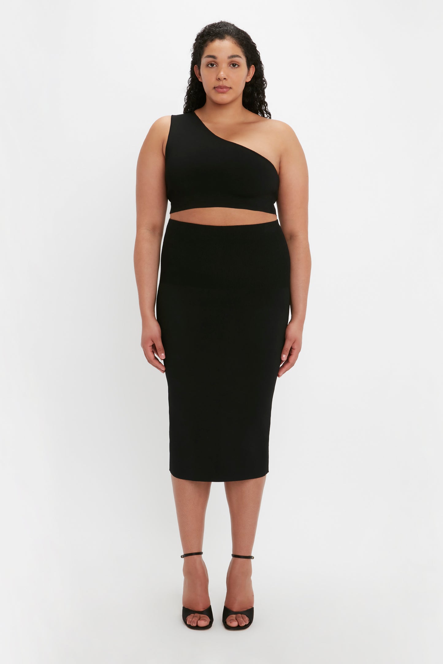 A person stands facing forward wearing the VB Body One Shoulder Crop Top in Black by Victoria Beckham and a matching black midi skirt, complemented with black high-heeled shoes.