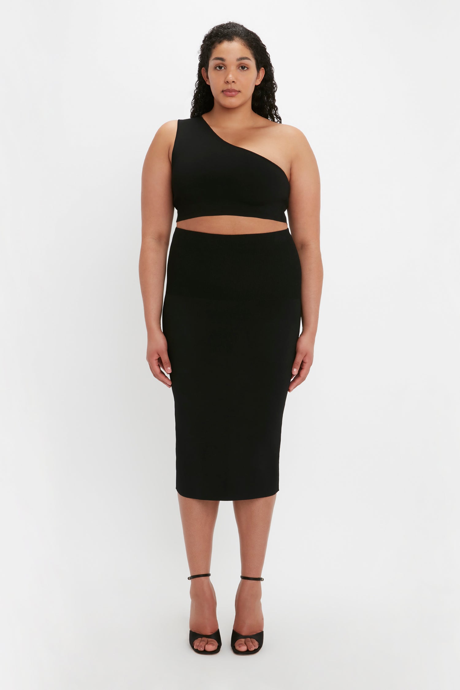 A person stands facing forward wearing the VB Body One Shoulder Crop Top in Black by Victoria Beckham and a matching black midi skirt, complemented with black high-heeled shoes.