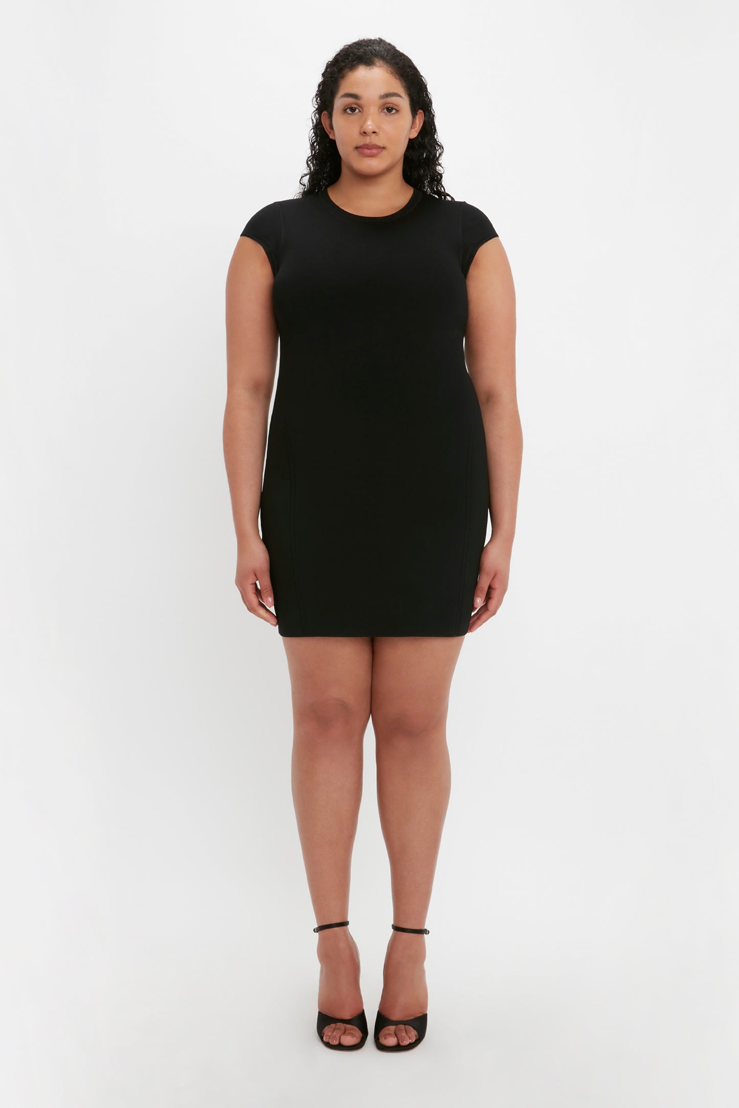 A woman with curly hair stands facing forward, wearing the VB Body Compact Cap Sleeve Mini Dress in Black by Victoria Beckham and black high-heeled shoes against a plain white background.