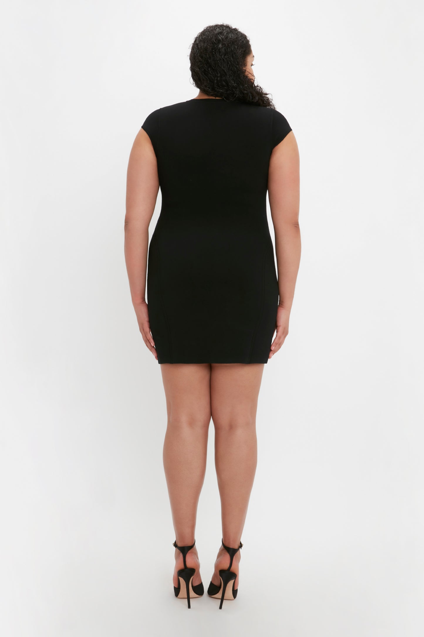 A person with curly hair, wearing the Victoria Beckham VB Body Compact Cap Sleeve Mini Dress in Black and high heels, stands facing away from the camera against a plain white background.