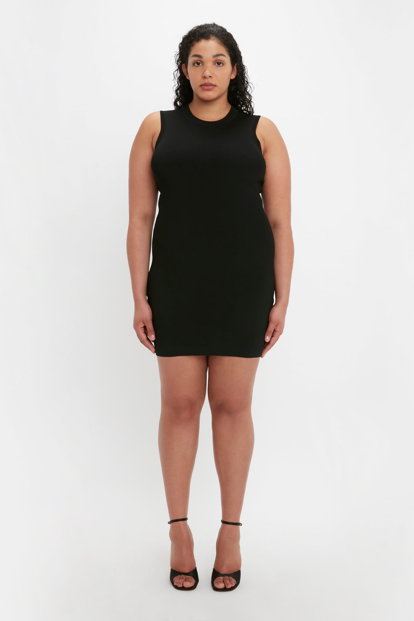 A woman with curly hair stands against a plain backdrop wearing the VB Body Mini Dress in Black by Victoria Beckham and black strappy heels.