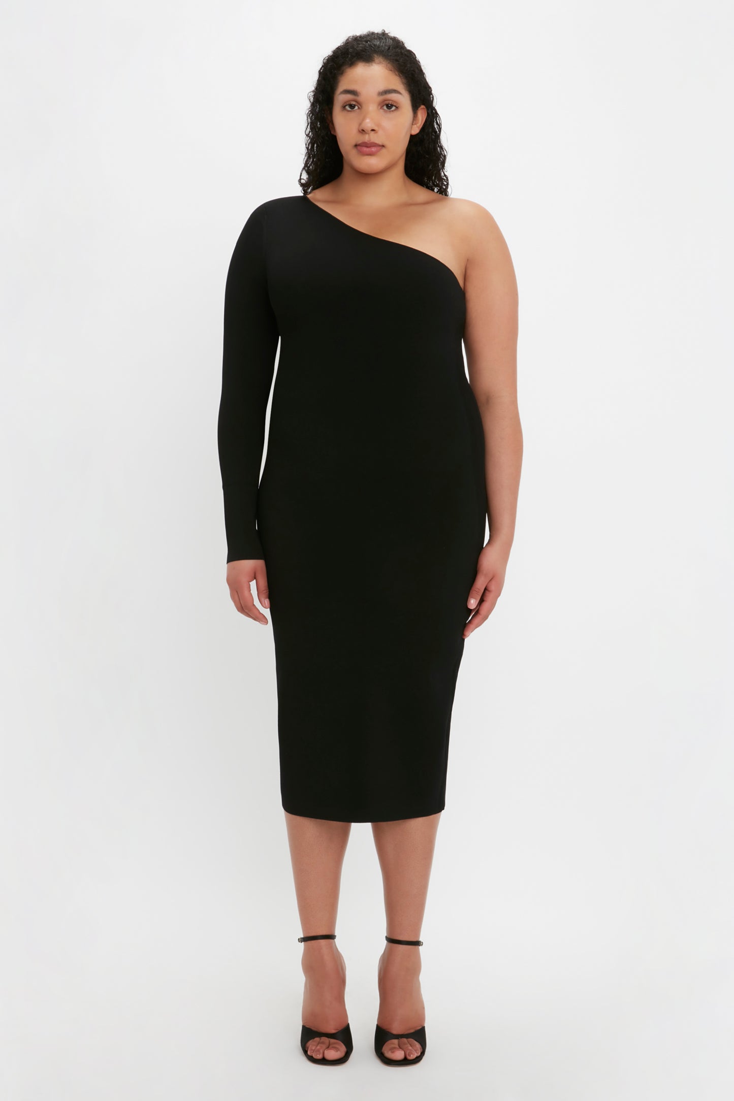 A woman stands facing forward, wearing the VB Body One Shoulder Midi Dress in Black by Victoria Beckham and black heels against a plain white background.