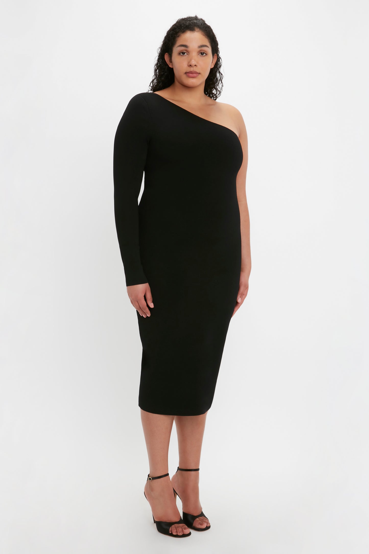 A woman stands against a plain background wearing the Victoria Beckham VB Body One Shoulder Midi Dress in Black paired with black heels.