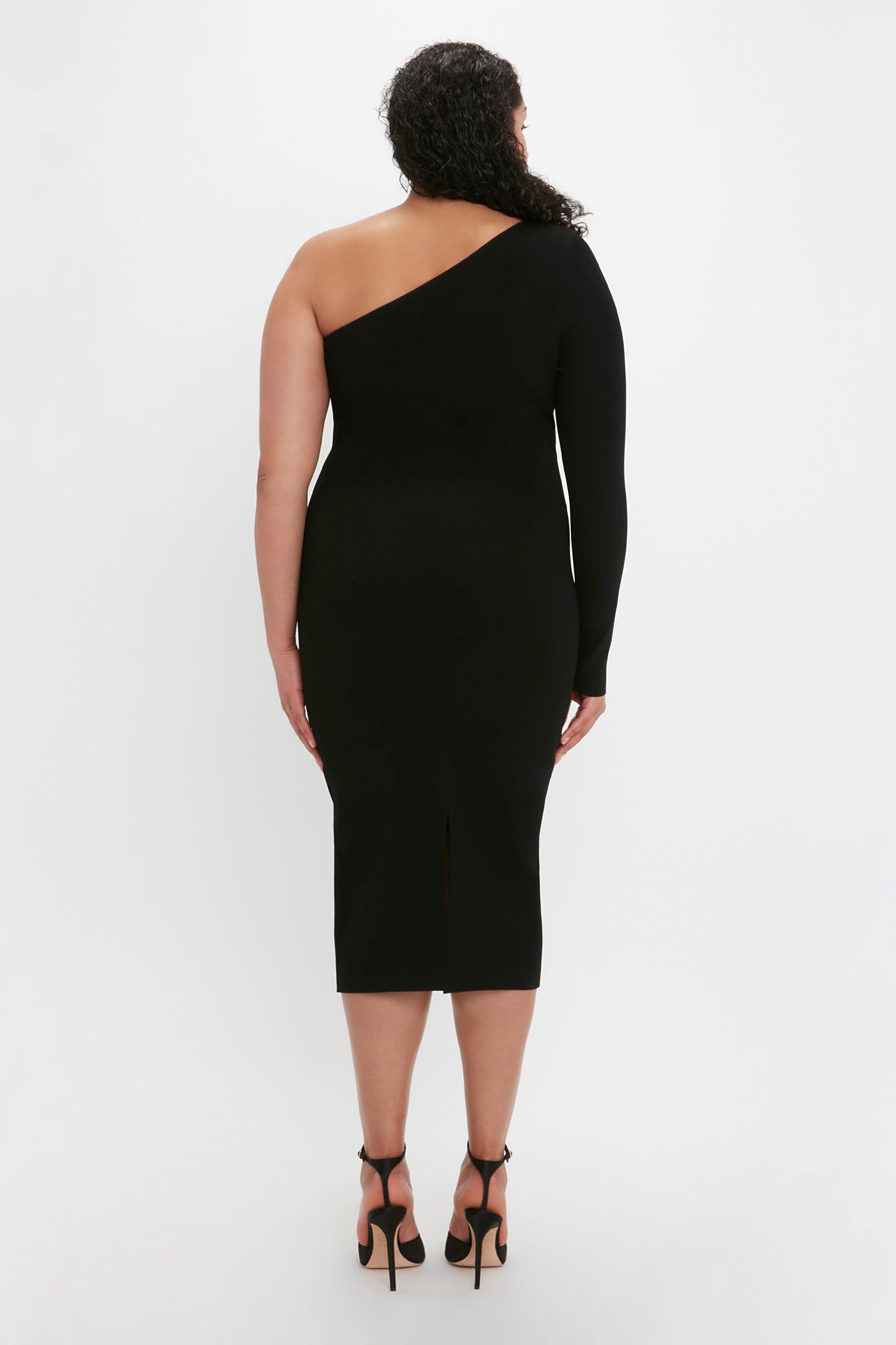 A person with curly hair stands facing away, wearing the VB Body One Shoulder Midi Dress in Black by Victoria Beckham and high-heeled shoes.