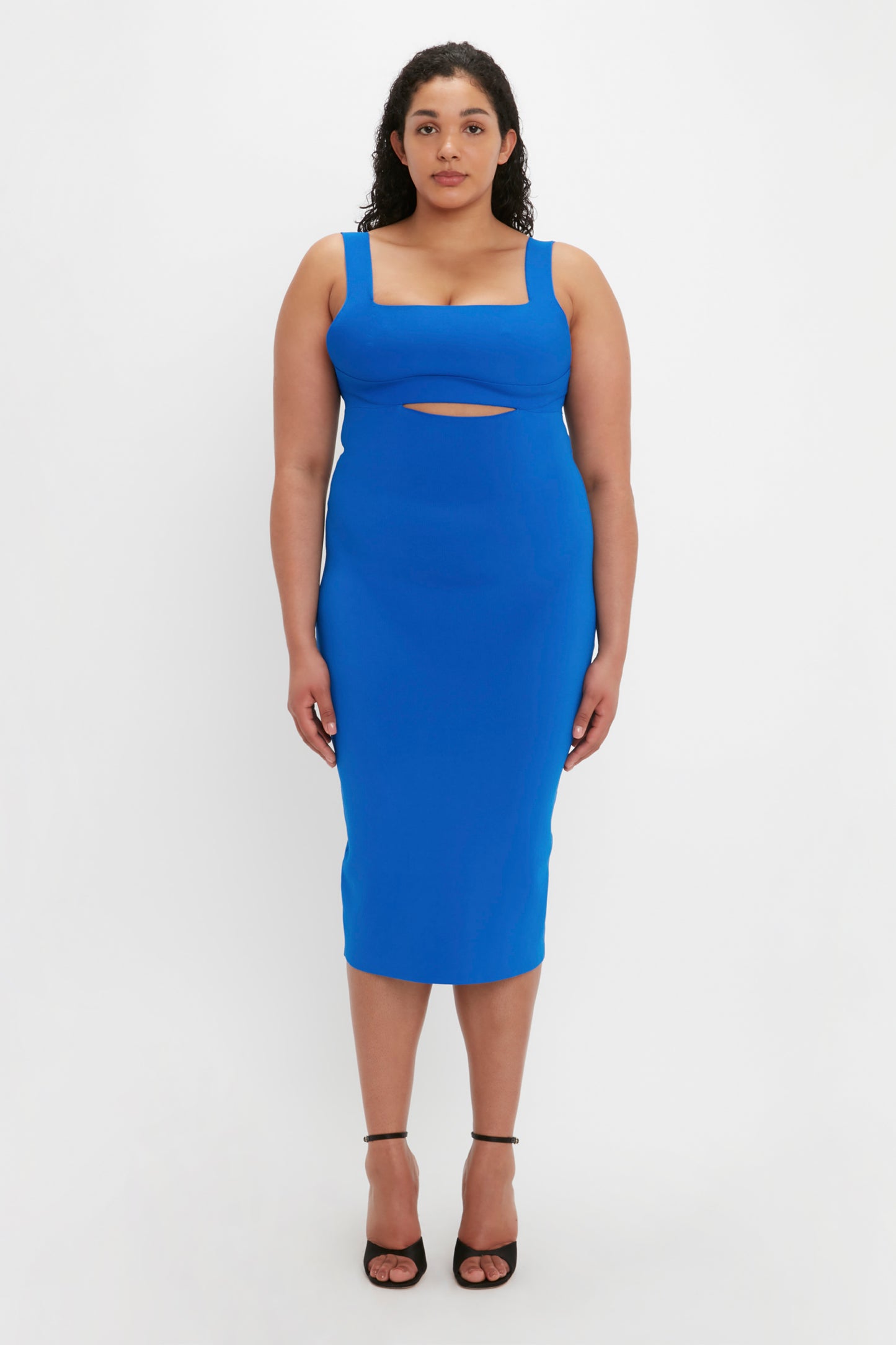 A person wearing the VB Body Cut Out Midi Dress in Cobalt by Victoria Beckham stands against a plain white background. They are also wearing black strappy heels.