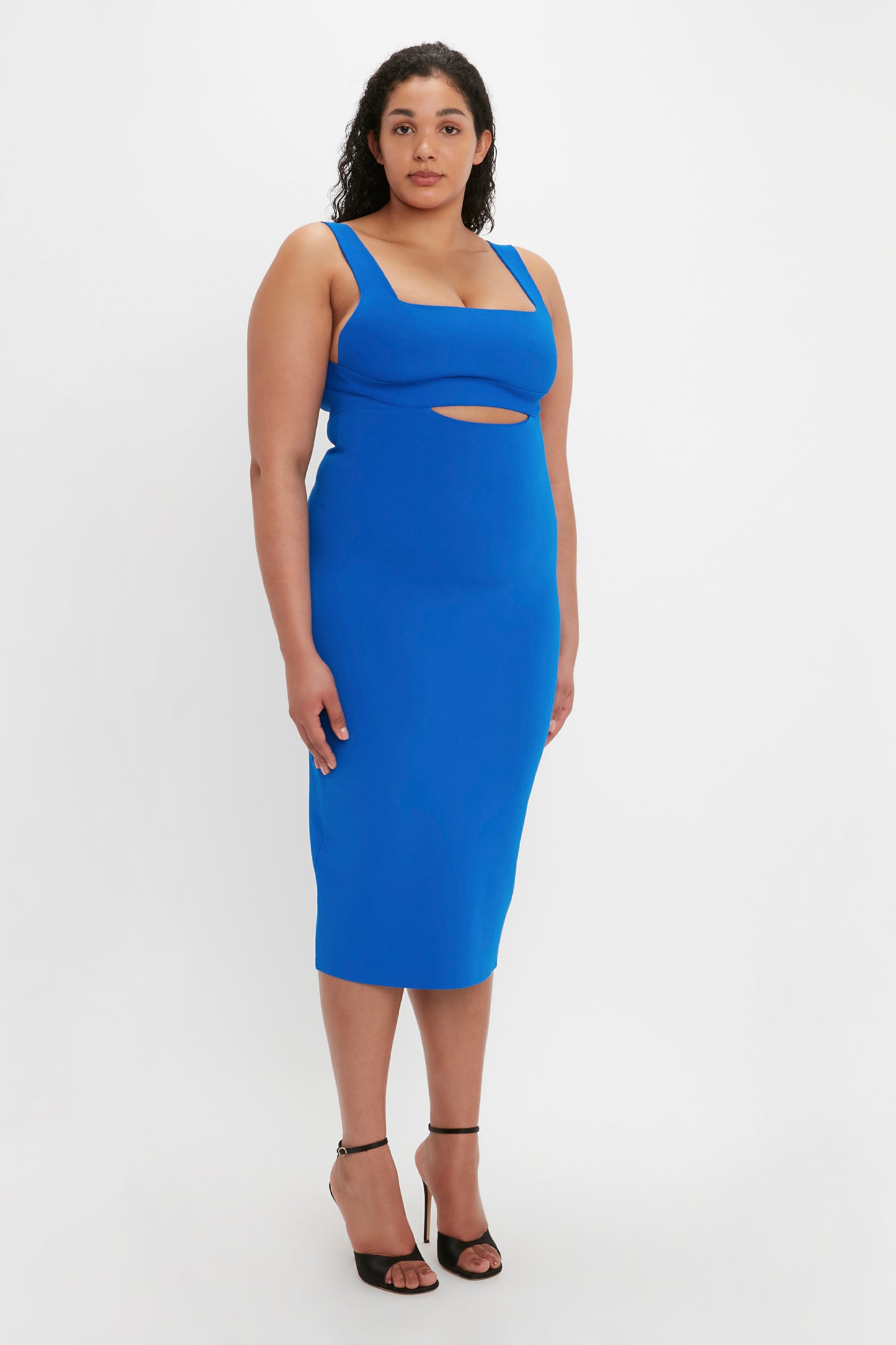 A woman stands against a white background wearing the VB Body Cut Out Midi Dress in Cobalt from Victoria Beckham, paired with black high-heeled shoes.