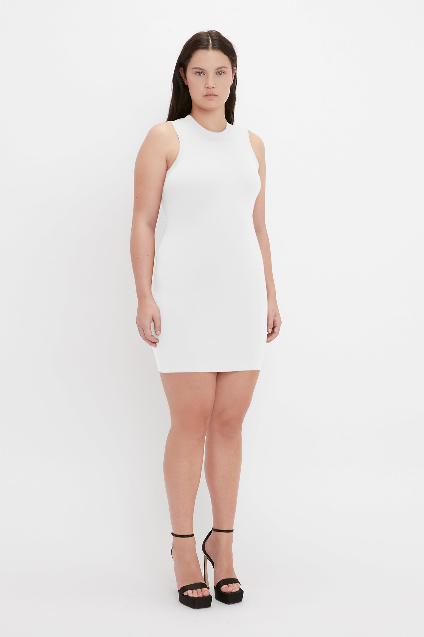 A woman standing against a plain white background, wearing the VB Body Mini Dress in White by Victoria Beckham and black high heels. She has long, dark hair and a neutral expression.