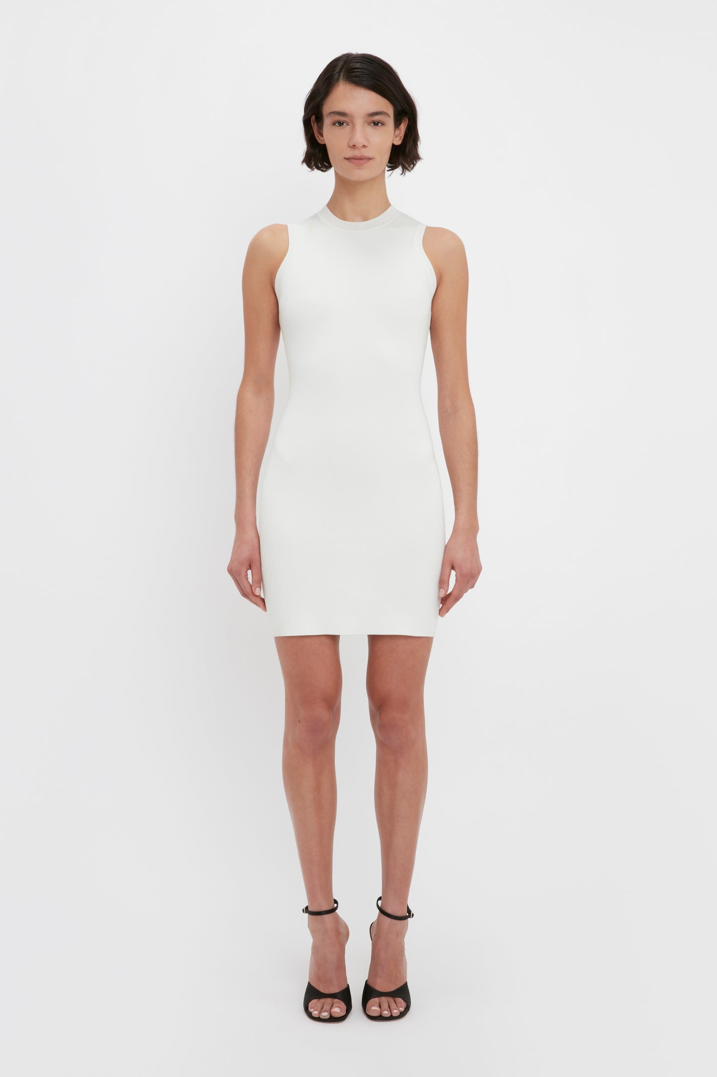 A person stands against a white background wearing the VB Body Mini Dress in White by Victoria Beckham, paired with black high-heeled sandals.