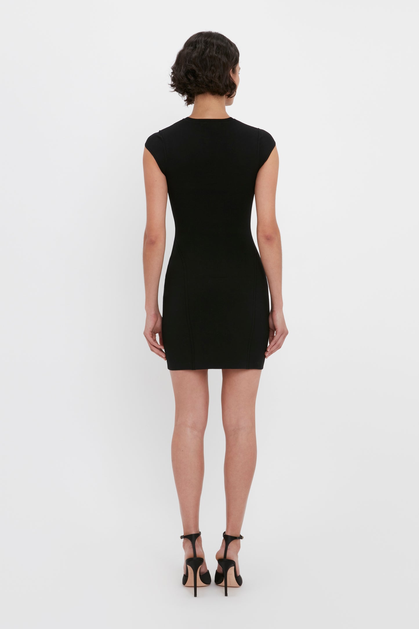 A person with short, curly hair is standing with their back to the camera, wearing the Victoria Beckham VB Body Compact Cap Sleeve Mini Dress in Black and black high heels.