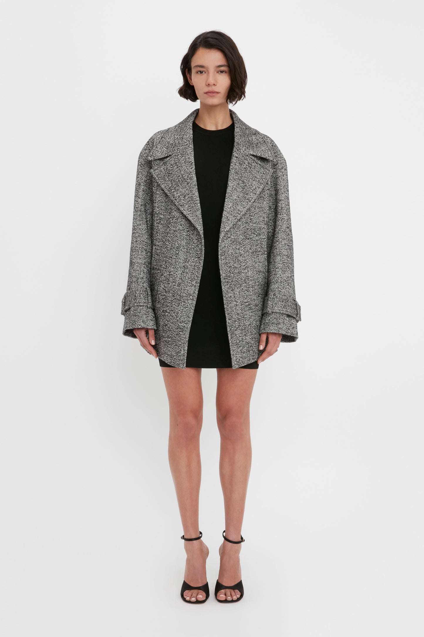 A person stands facing forward wearing a grey oversized coat over the VB Body Mini Dress in Black by Victoria Beckham with black high-heeled sandals.