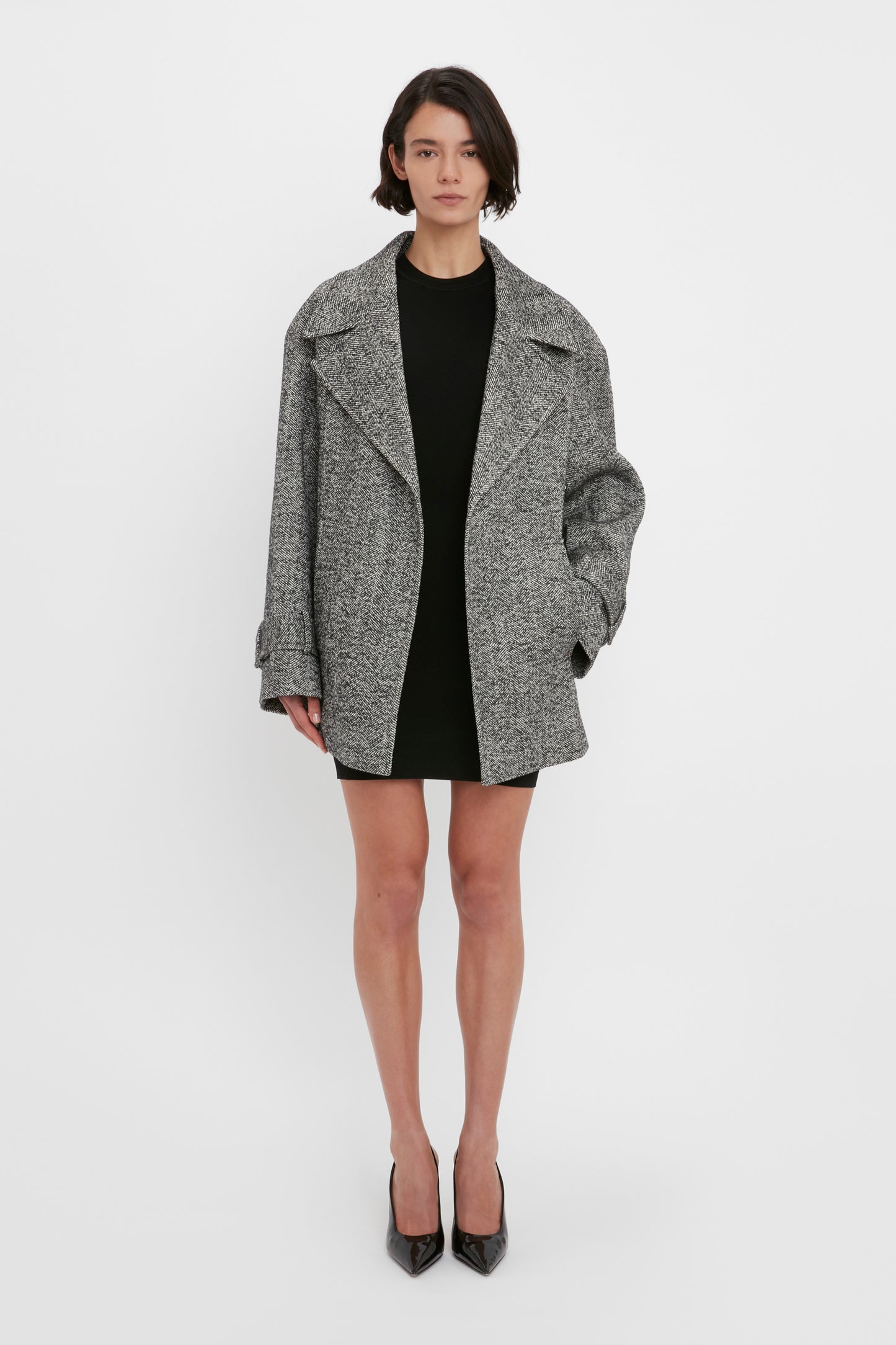 Person standing against a white background wearing a grey oversized coat, Victoria Beckham's VB Body Mini Dress in Black, and black heels.