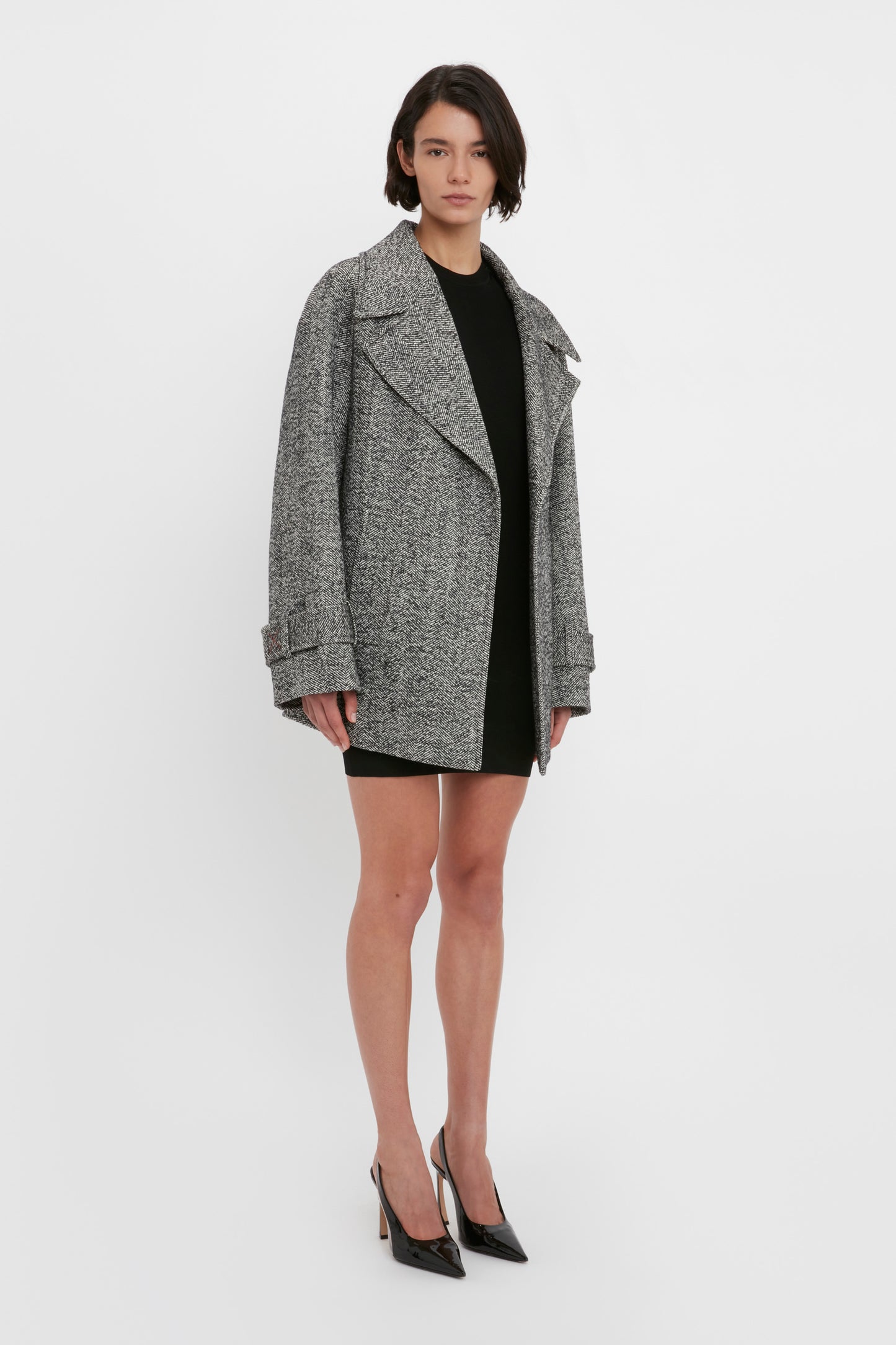 A person stands against a white background wearing an oversized gray coat, the VB Body Mini Dress in Black by Victoria Beckham, and black high heels.