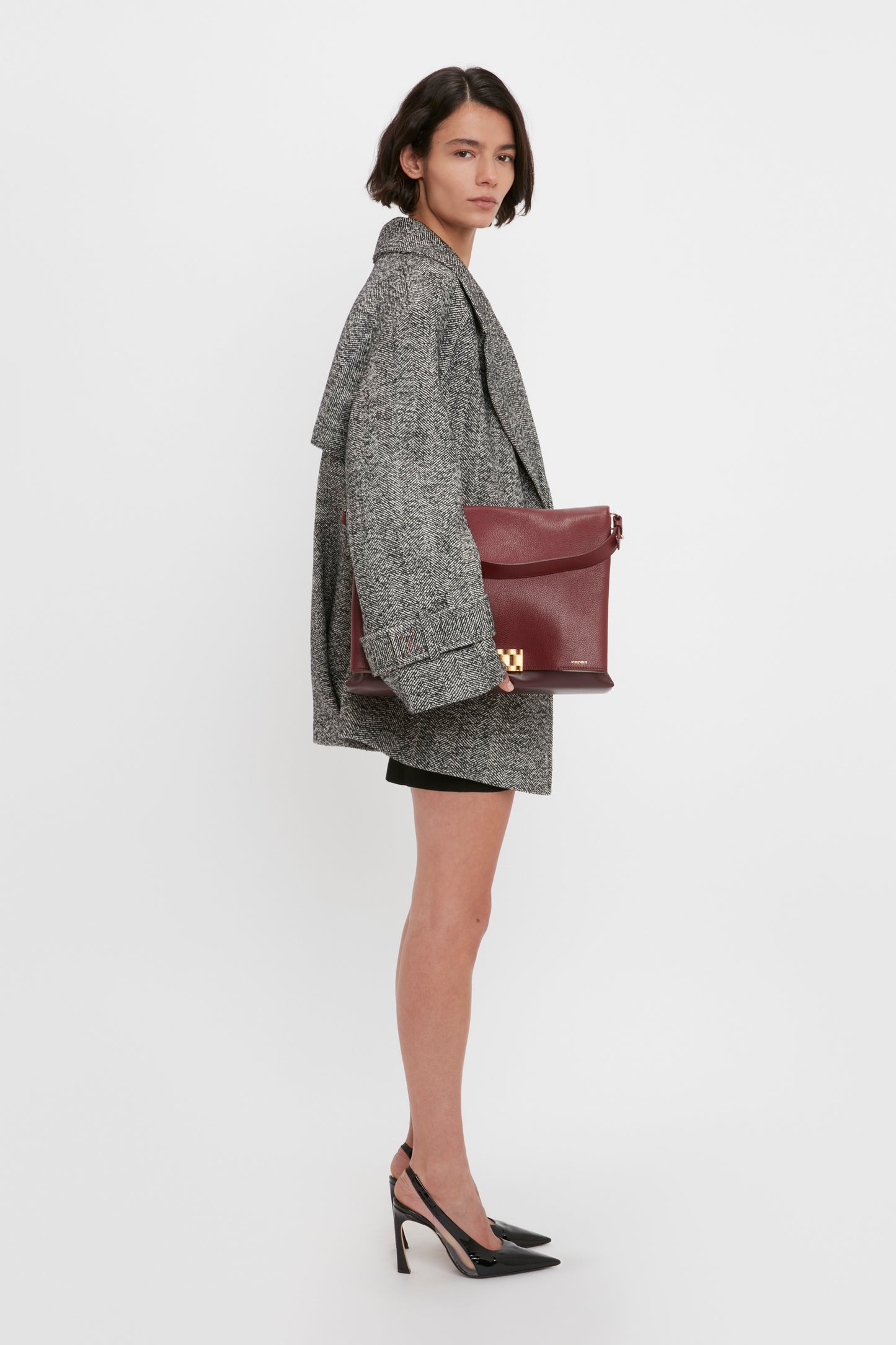 A woman stands sideways in a studio wearing the Victoria Beckham VB Body Mini Dress in Black, black high heels, and holding a large maroon clutch bag.