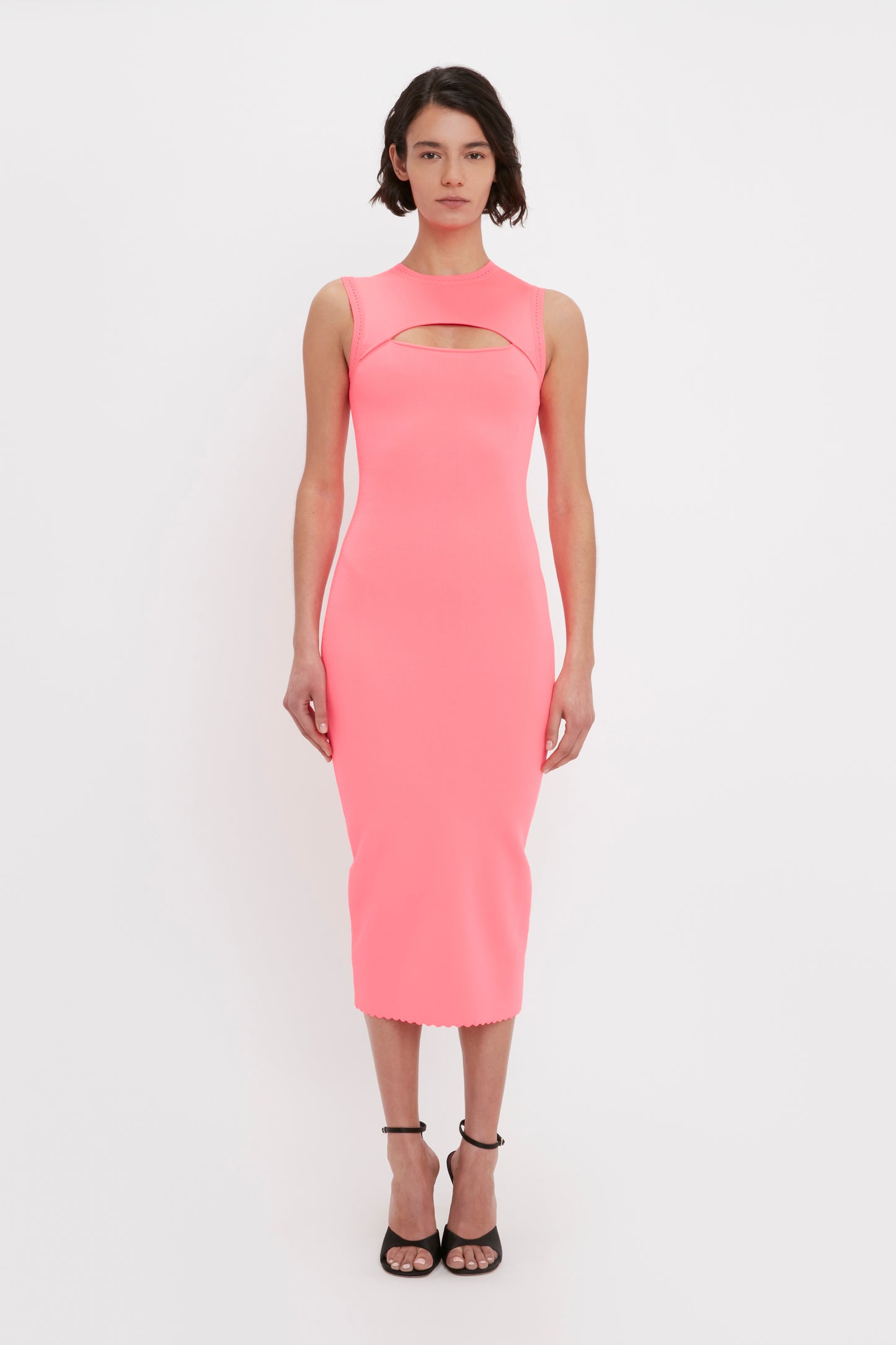 A person standing against a plain background, wearing the VB Body Slash Front Midi Dress in Pink by Victoria Beckham, paired with black high-heeled sandals.