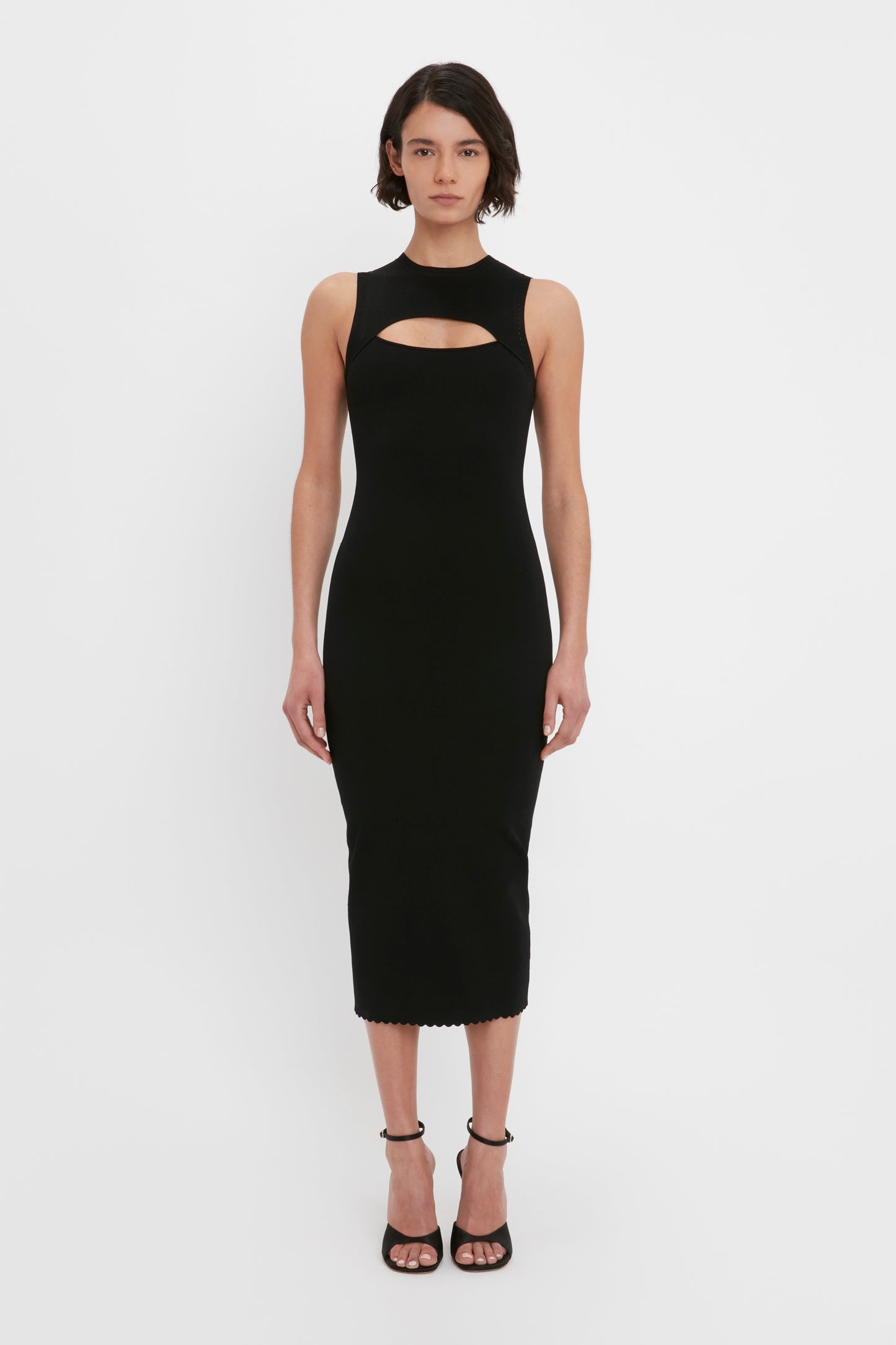 A woman with short dark hair is wearing the VB Body Cut Out Midi Dress in black by Victoria Beckham, paired with black high-heeled sandals, standing against a plain white background.