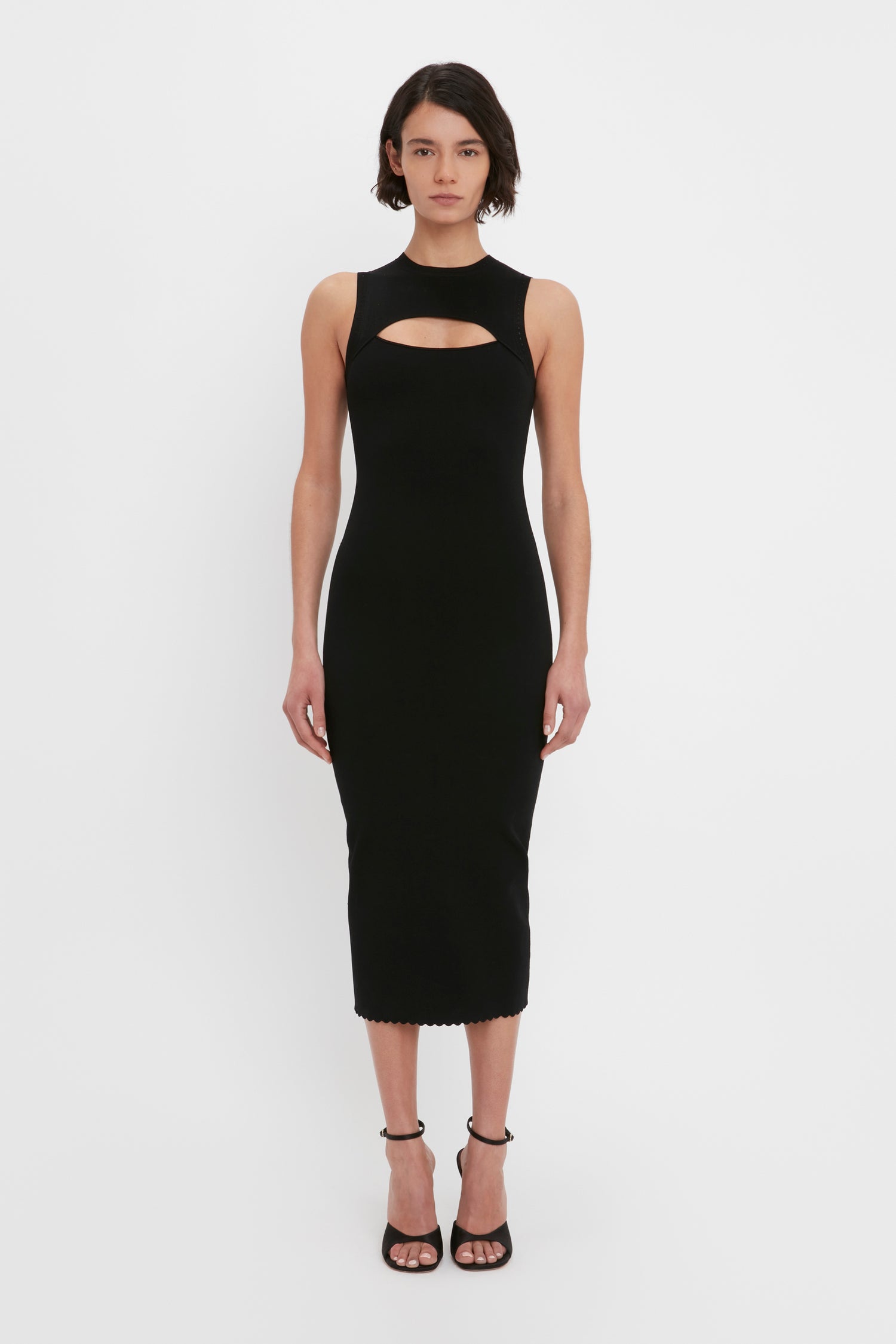 A woman with short dark hair is wearing the VB Body Cut Out Midi Dress in black by Victoria Beckham, paired with black high-heeled sandals, standing against a plain white background.