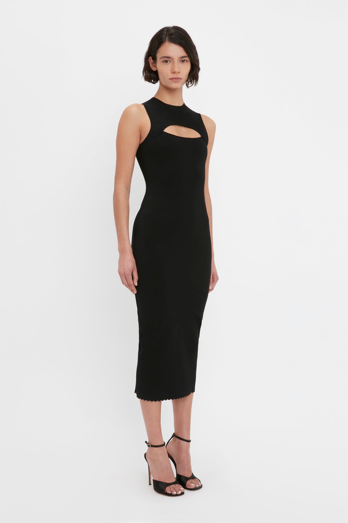 A woman stands against a white background wearing the VB Body Cut Out Midi Dress in Black by Victoria Beckham, paired with black high-heeled sandals.