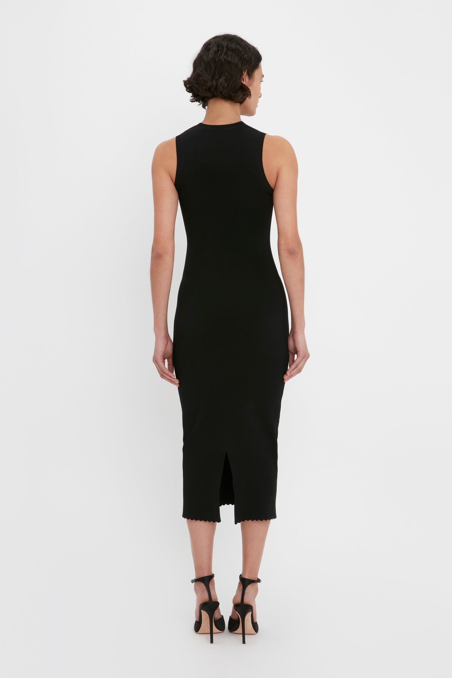 A person with short dark hair is shown from the back, wearing a sleeveless VB Body Cut Out Midi Dress in Black by Victoria Beckham and black high-heeled sandals.