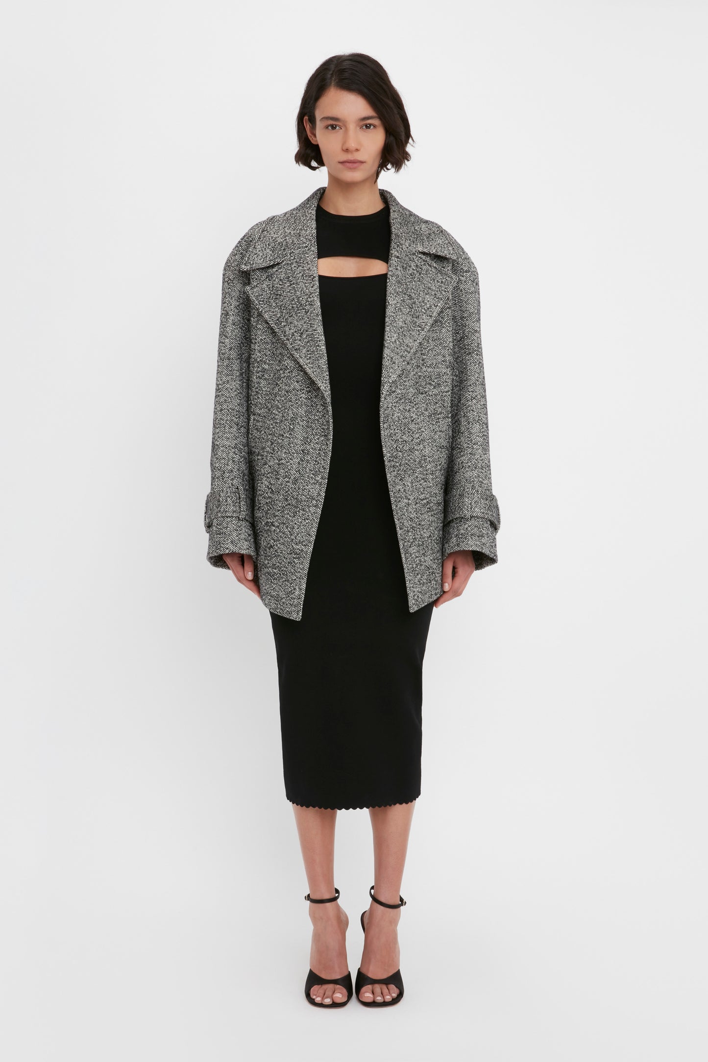 An individual wearing the Victoria Beckham VB Body Cut Out Midi Dress in Black, styled with a grey oversized coat and black heeled sandals, stands against a plain white background.