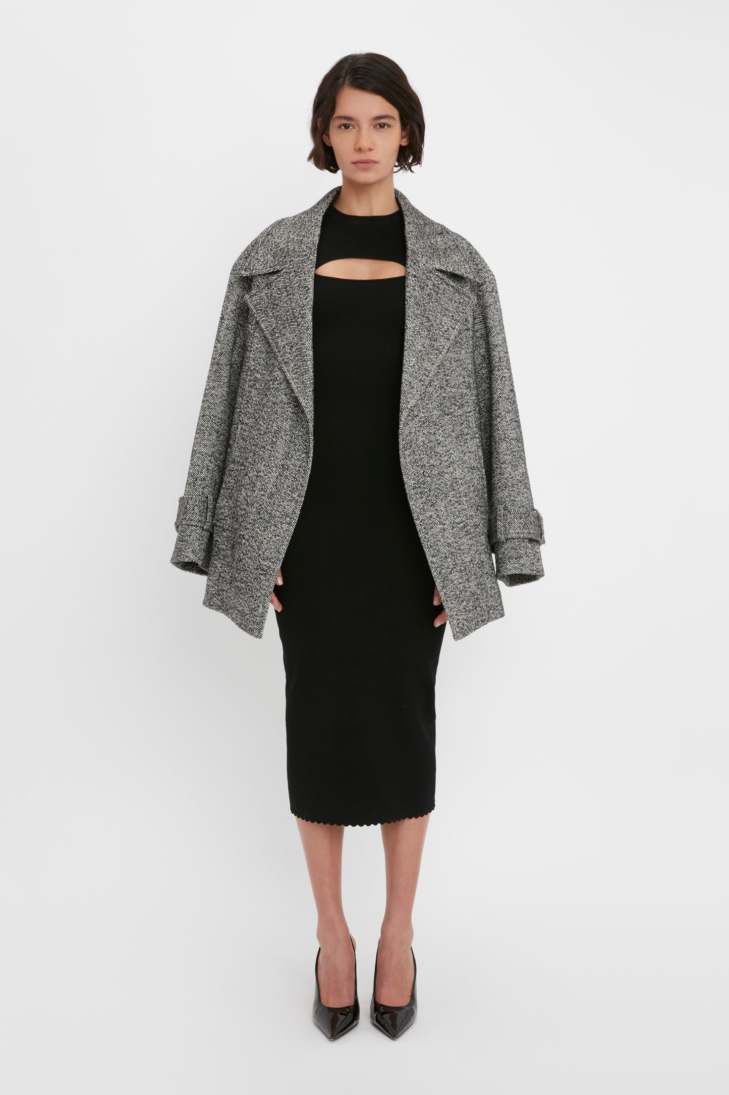 A person stands against a plain background wearing a gray coat over the VB Body Cut Out Midi Dress in Black by Victoria Beckham and black heels.