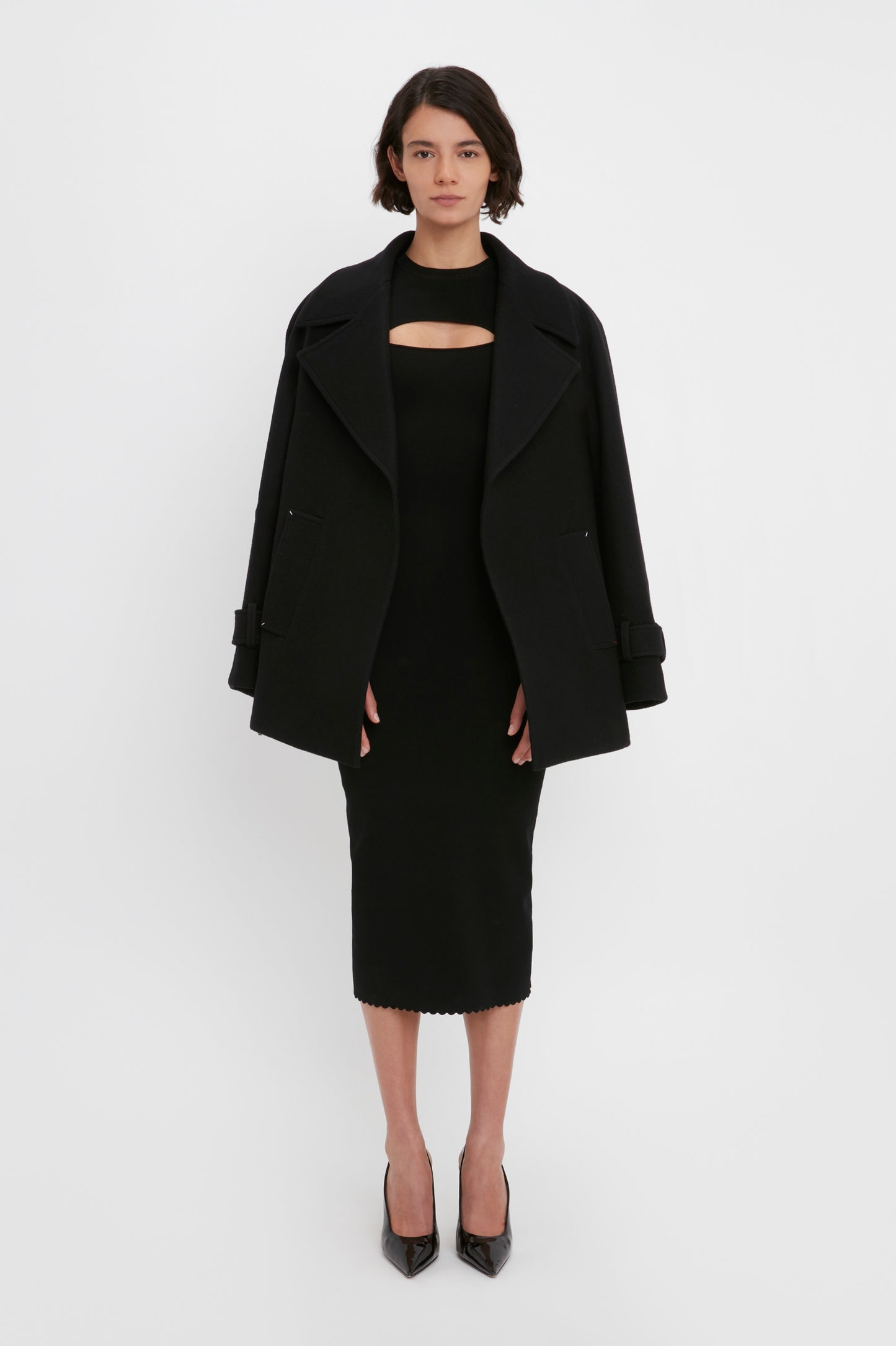 A person stands against a white background, wearing the VB Body Cut Out Midi Dress in Black from Victoria Beckham with a black jacket draped over their shoulders. They are also wearing black heels.