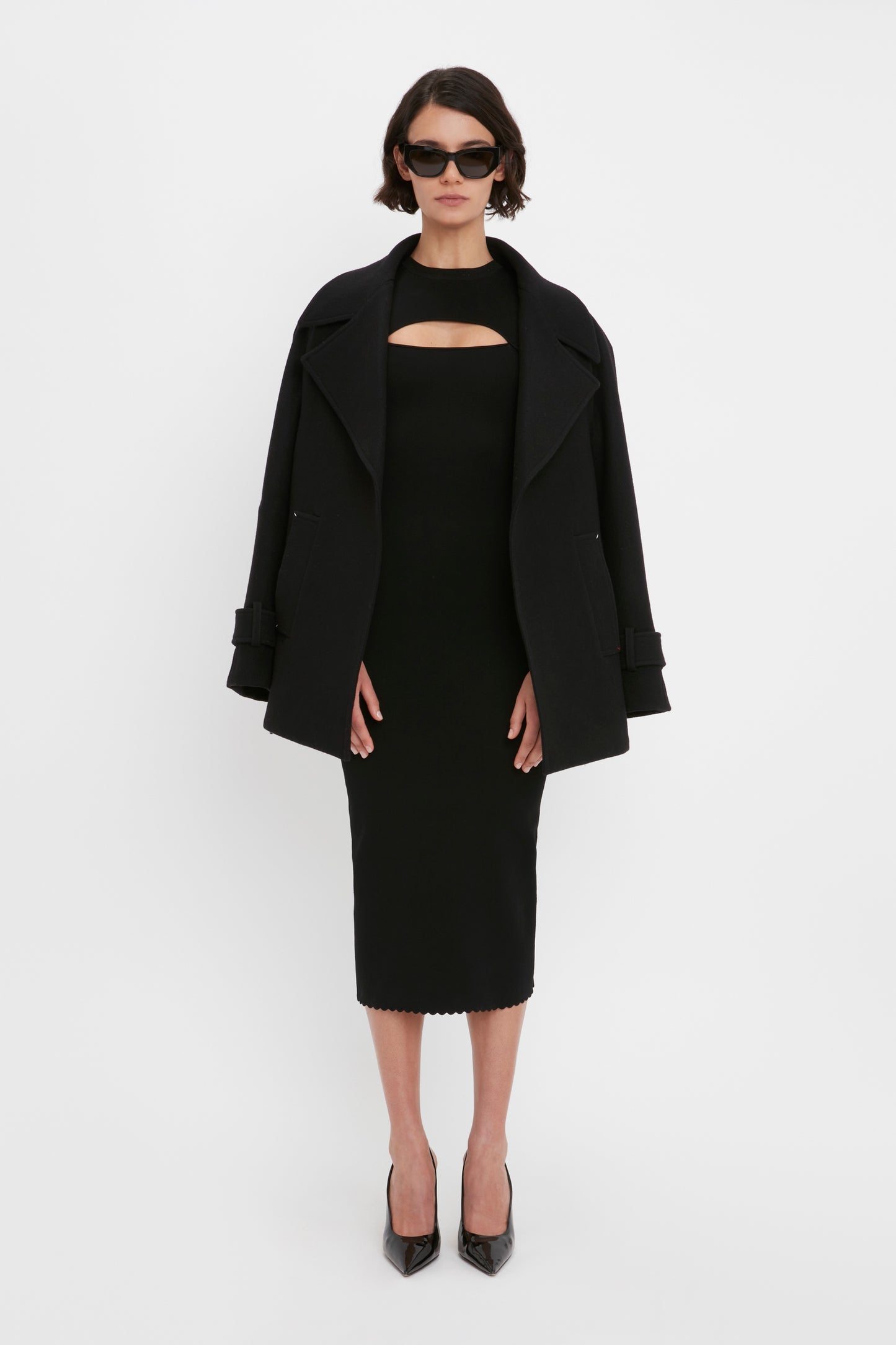 A woman in a VB Body Cut Out Midi Dress by Victoria Beckham, paired with a black oversized coat, black high heels, and sunglasses, standing against a plain white background.