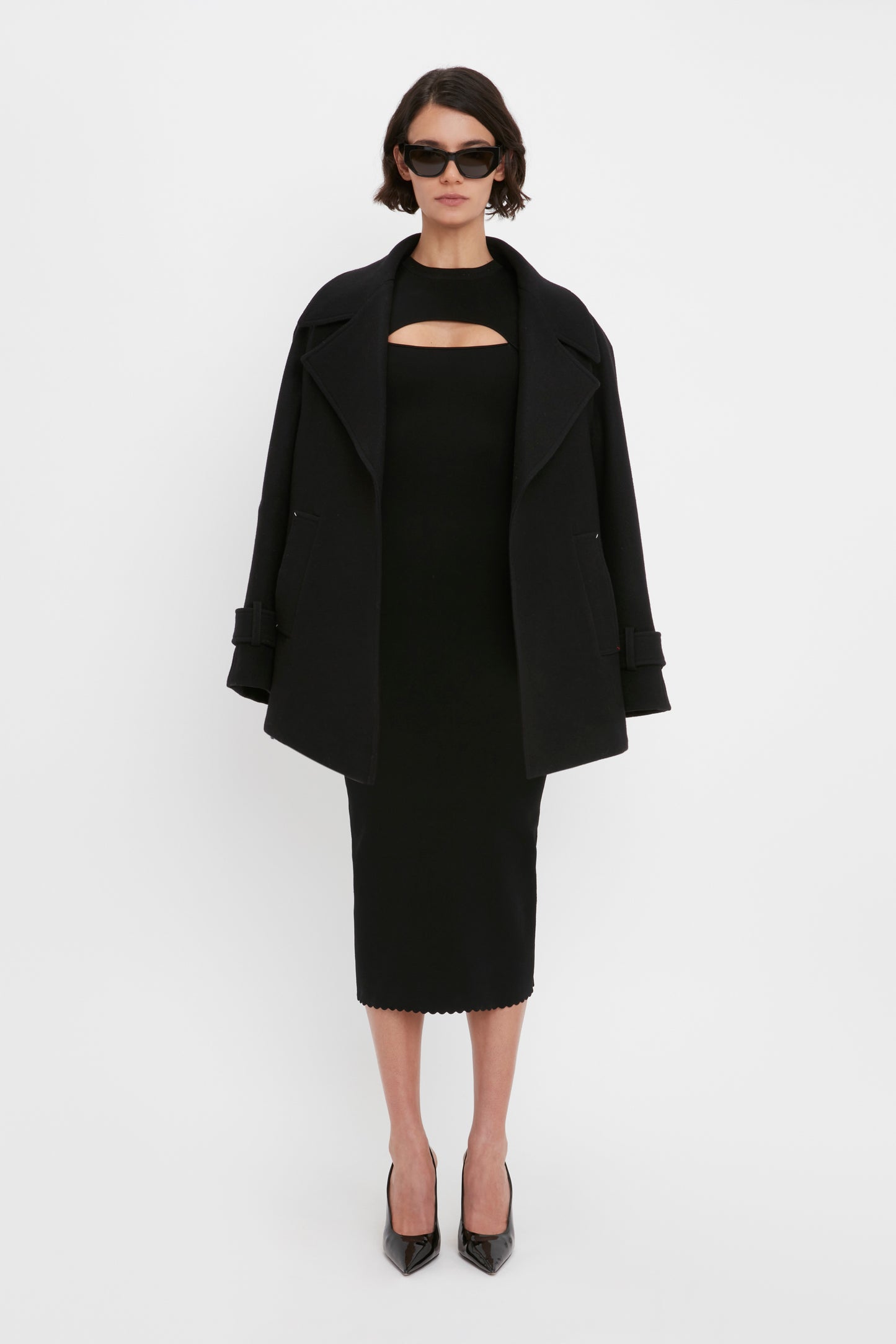 A person is standing against a white background, wearing the VB Body Cut Out Midi Dress in Black by Victoria Beckham, complemented with a black coat, black high-heeled shoes, and sunglasses.