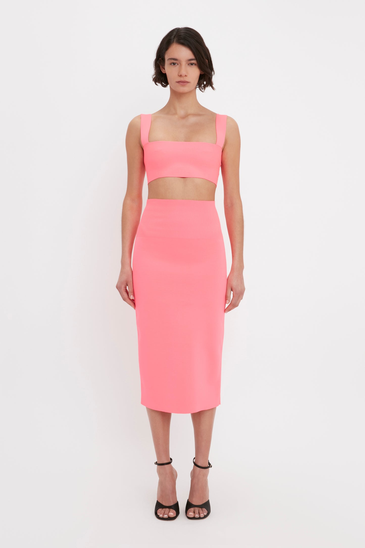 A person stands wearing a pink crop top and the VB Body Fitted Midi Skirt in Pink by Victoria Beckham, paired with black high-heeled sandals. They have short dark hair and are facing forward against a plain white background.