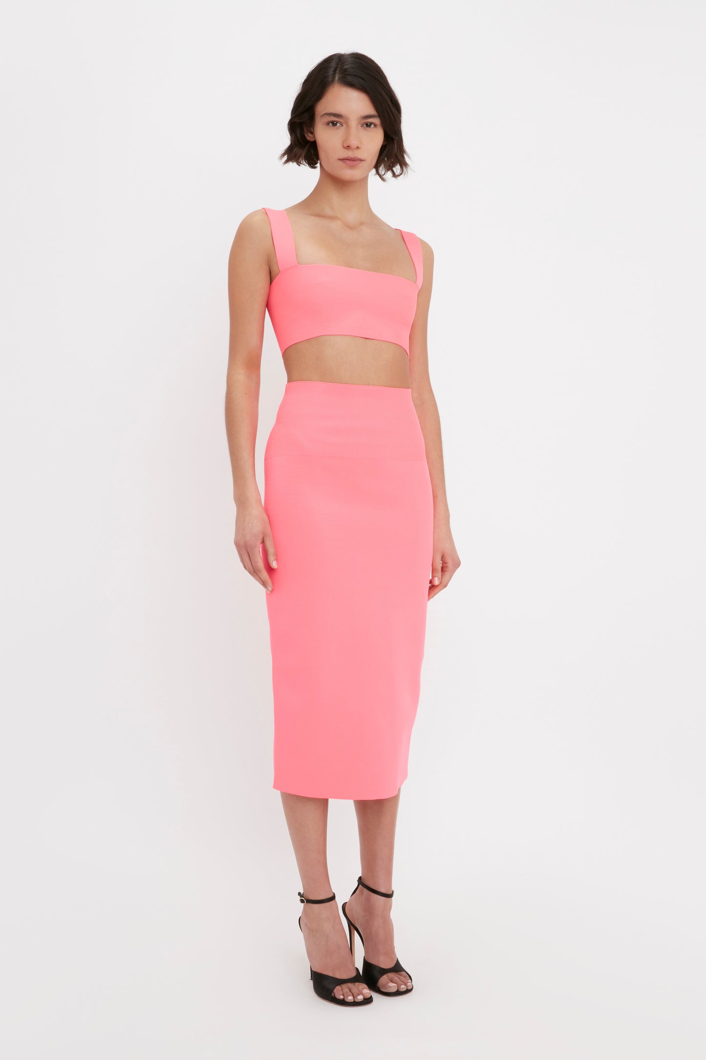 A woman stands against a plain background wearing the VB Body Strap Bandeau Top in Pink by Victoria Beckham, paired with a matching high-waisted skirt and black high-heeled sandals.