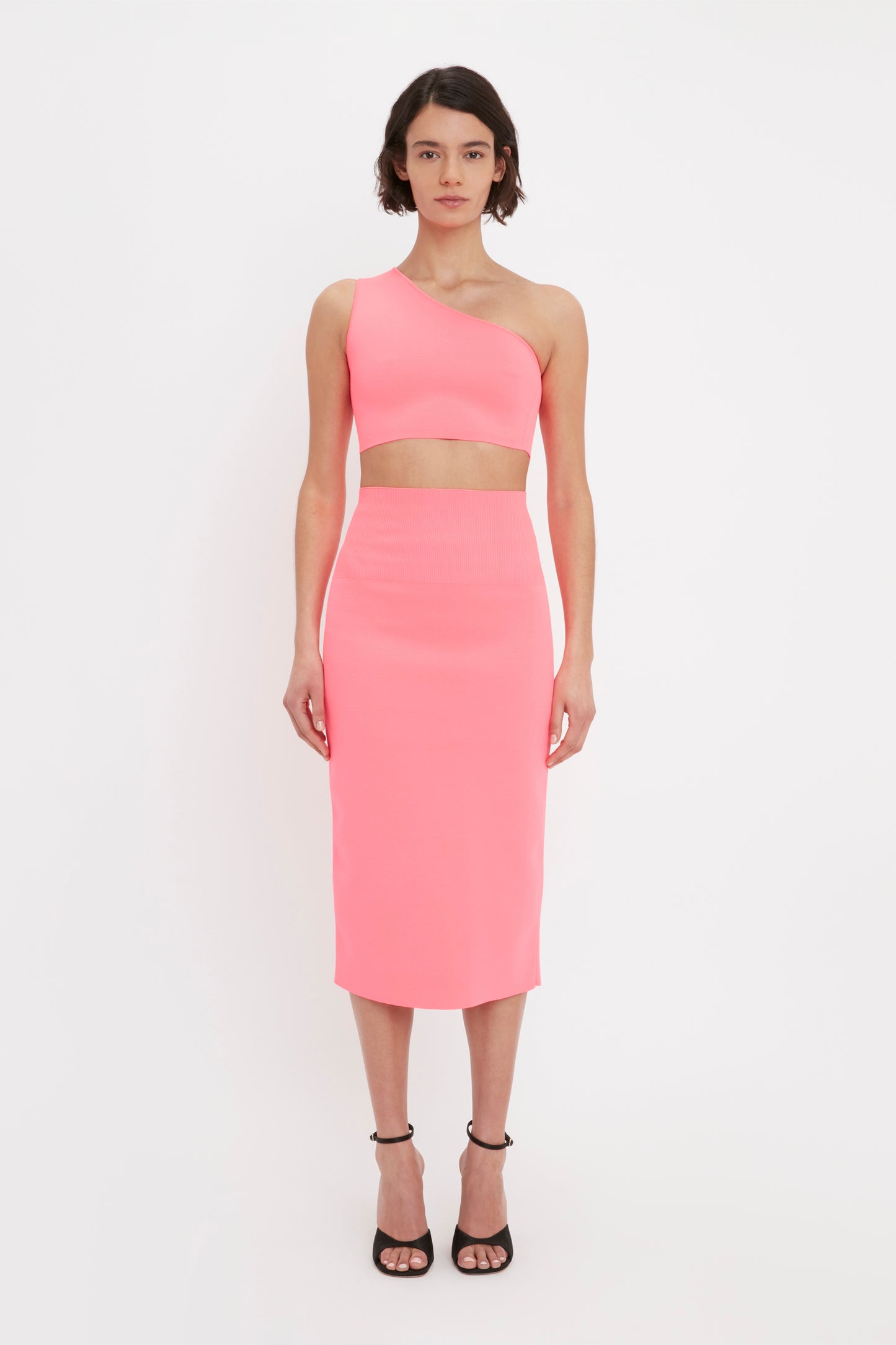 A woman wears the Victoria Beckham VB Body One Shoulder Crop Top in Pink along with a matching midi skirt and black heels, posing against a plain white background.