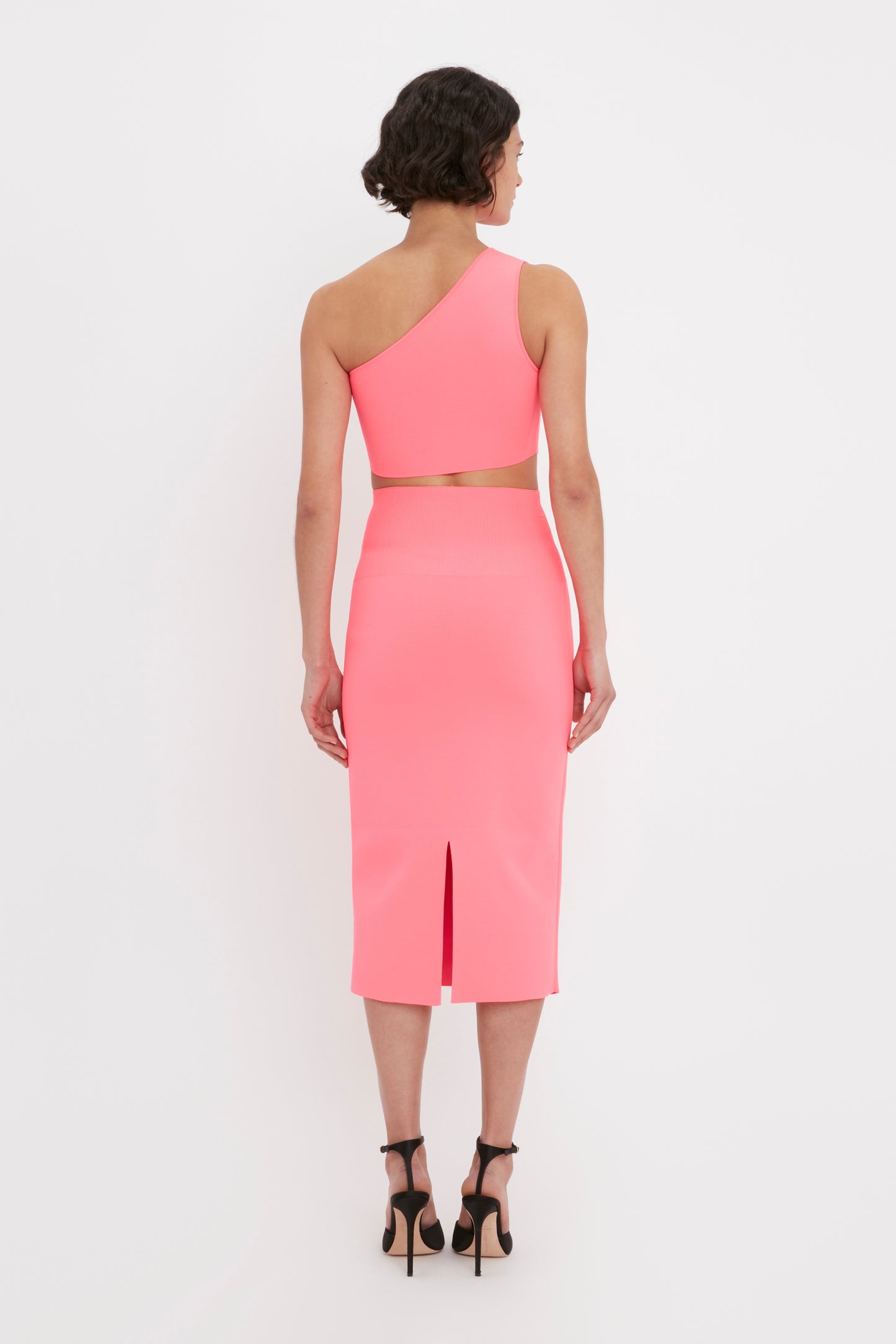A woman with short dark hair, wearing the Victoria Beckham VB Body One Shoulder Crop Top in pink and black high heels, is standing facing away from the camera.