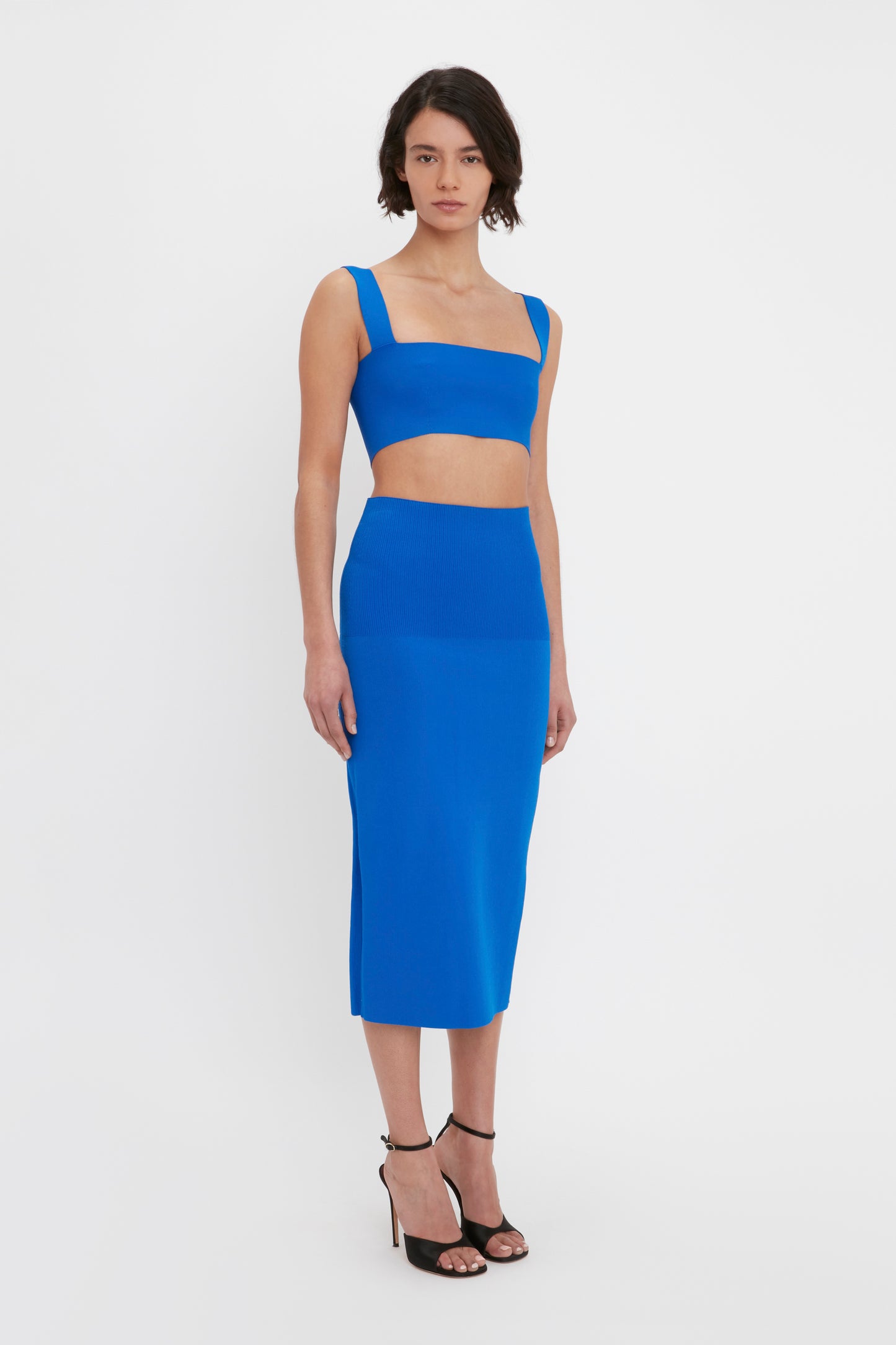 A person stands against a plain background, wearing a bright blue two-piece outfit from Victoria Beckham, which includes the VB Body Fitted Midi Skirt in Cobalt and a matching crop top, paired with black strappy heels.
