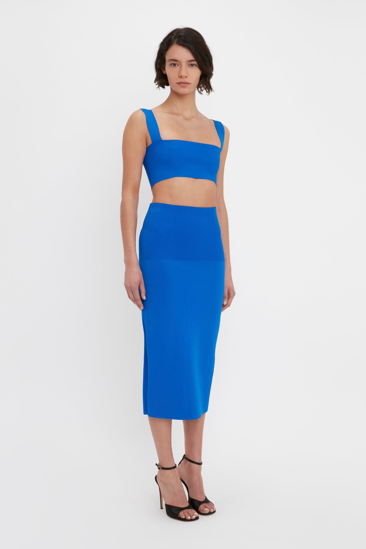 A person stands against a plain background, wearing a bright blue two-piece outfit from Victoria Beckham, which includes the VB Body Fitted Midi Skirt in Cobalt and a matching crop top, paired with black strappy heels.