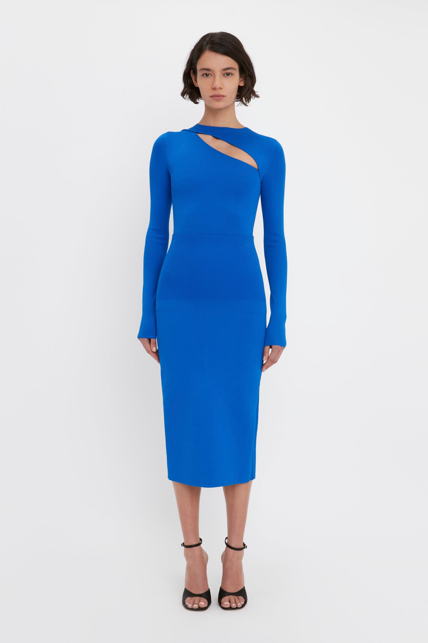 A woman is standing against a white background wearing the VB Body Front Slash Crew Neck Top in Cobalt from Victoria Beckham, paired with black strappy heels.