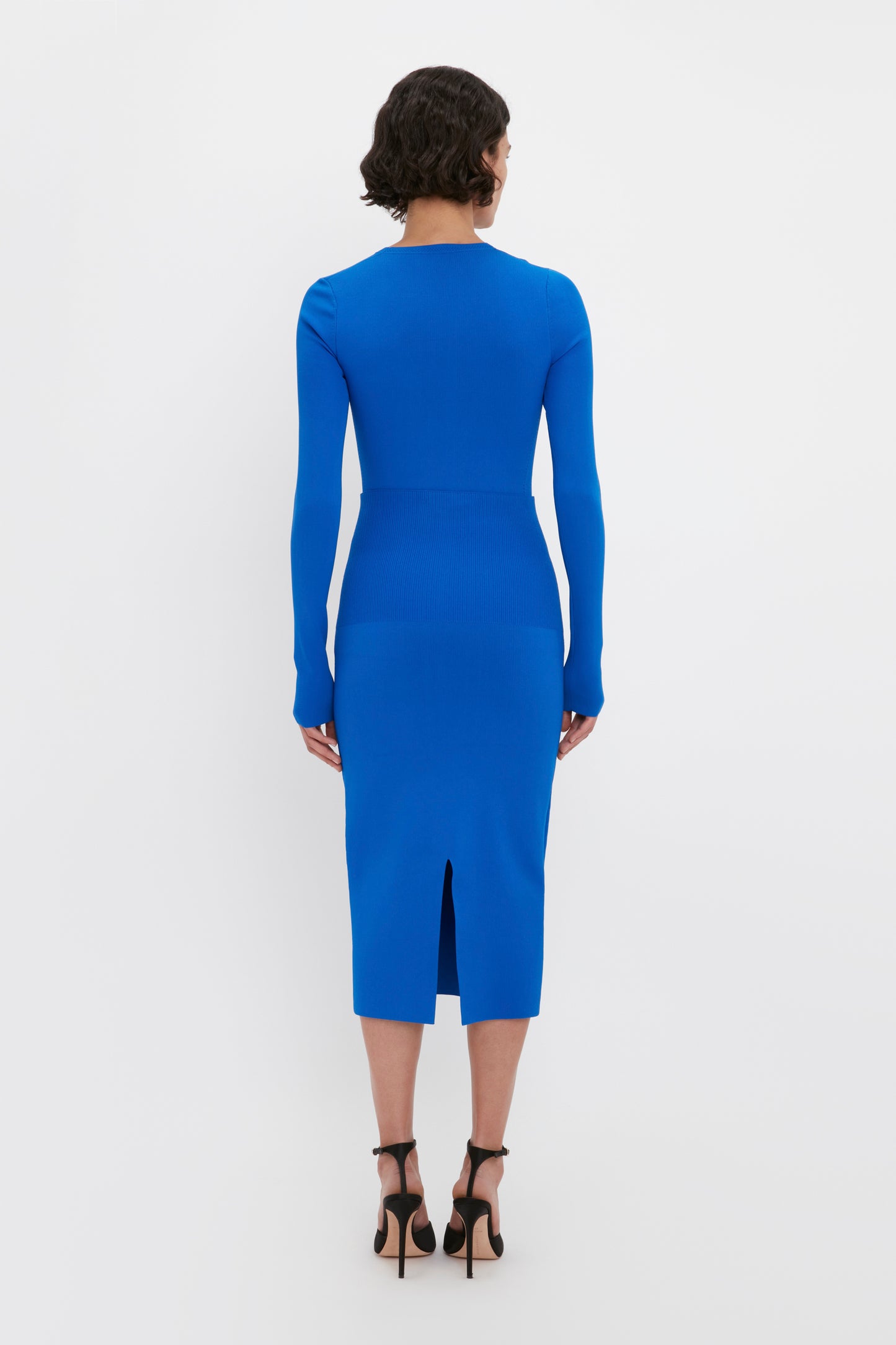 Person standing with back to camera, wearing a form-fitting Victoria Beckham Front Slash Crew Neck Top in Cobalt and black high heels.