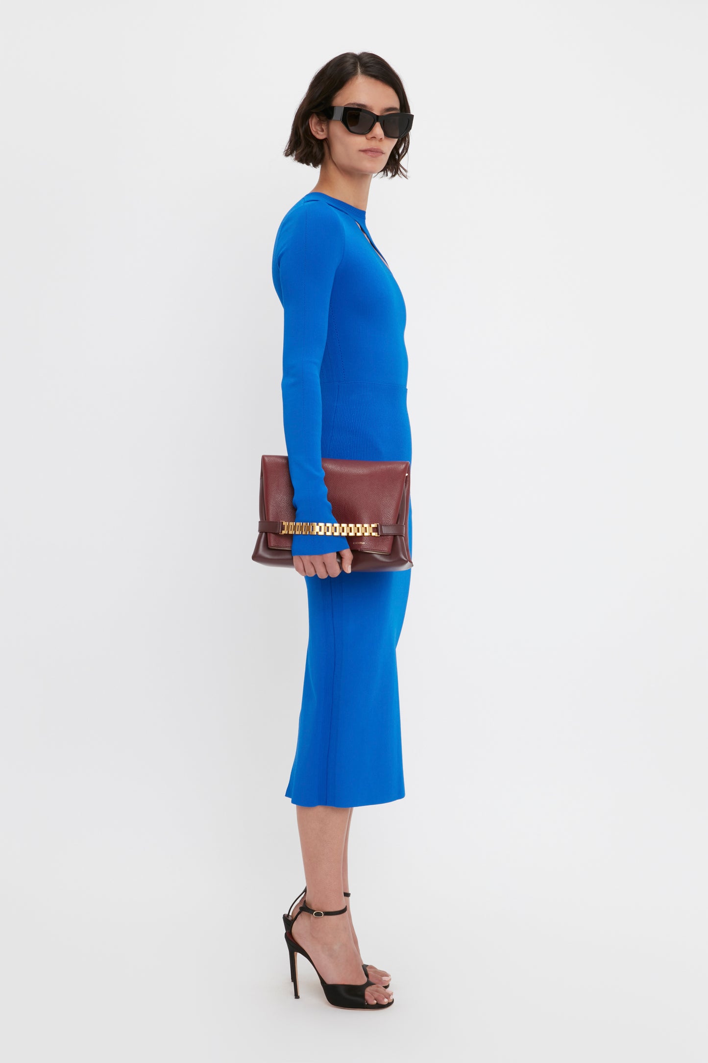 A person wearing the Victoria Beckham VB Body Front Slash Crew Neck Top in Cobalt and black high heels holds a brown clutch and wears dark sunglasses, standing against a plain white background.