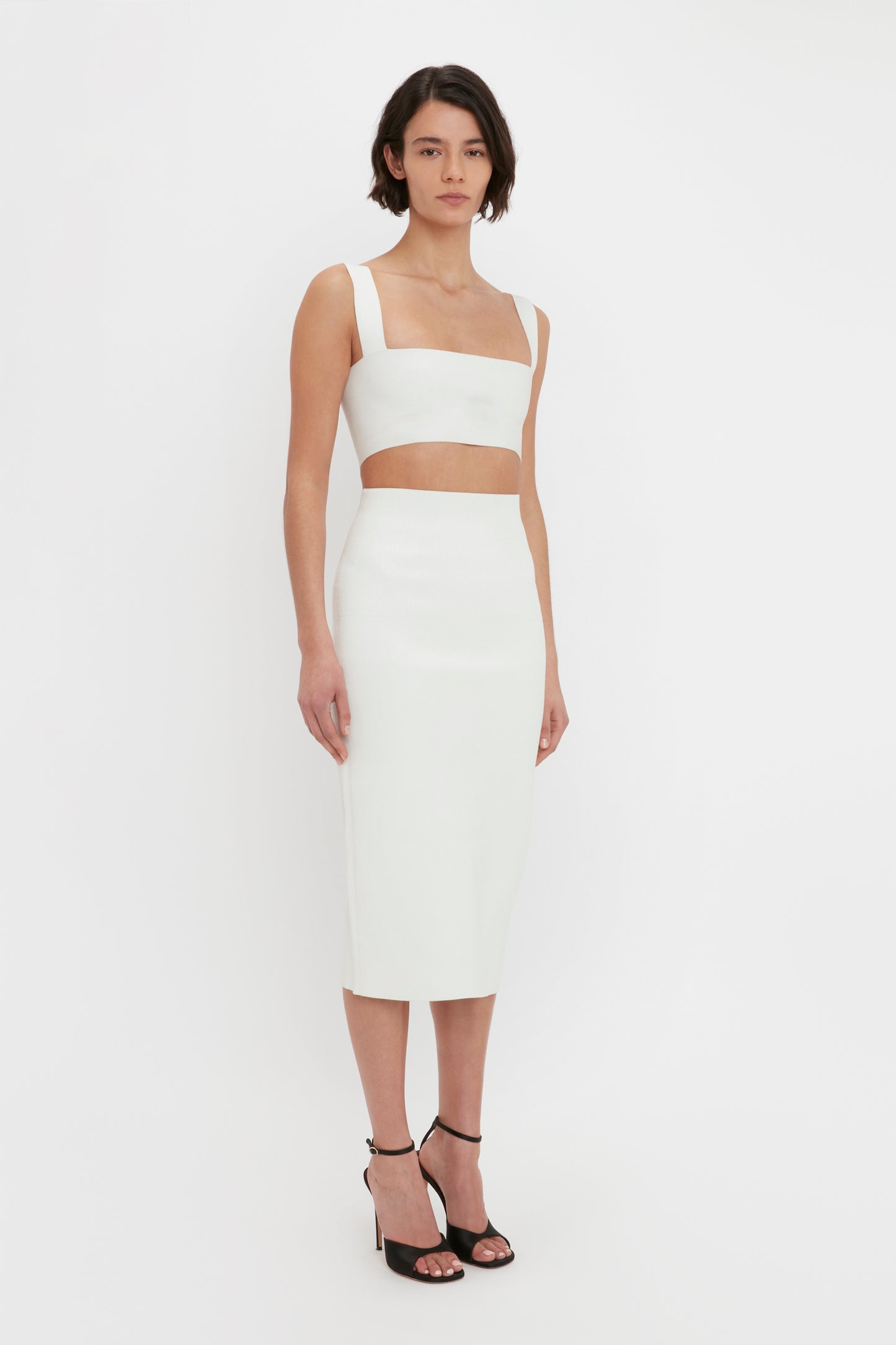 A woman standing against a white background, wearing the VB Body Strap Bandeau Top in White from Victoria Beckham and a coordinating high-waisted skirt, paired with black high-heeled shoes.
