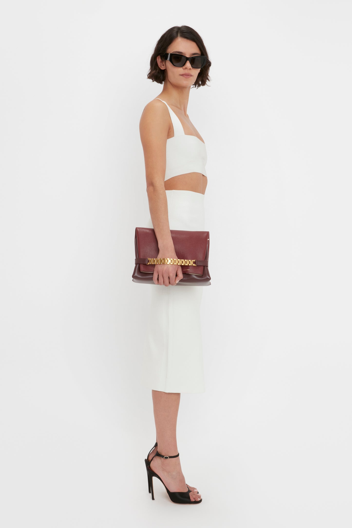 A woman wearing a Victoria Beckham VB Body Strap Bandeau Top in white and black high heels holds a maroon clutch bag. She is also sporting dark sunglasses.