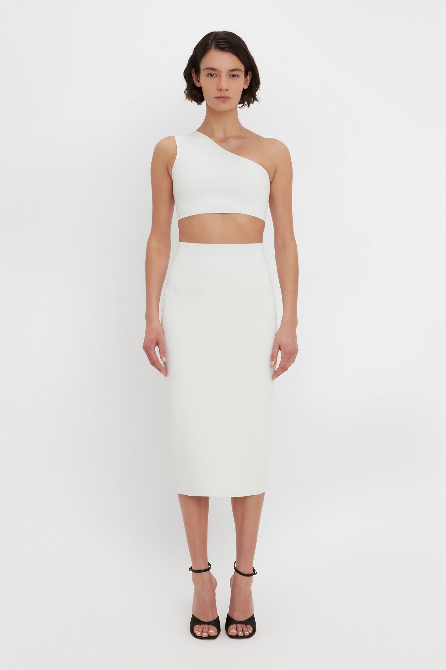 A person stands against a white background wearing a white, one-shoulder, midriff-baring top and the VB Body Fitted Midi Skirt in White by Victoria Beckham, paired with black high-heeled sandals.