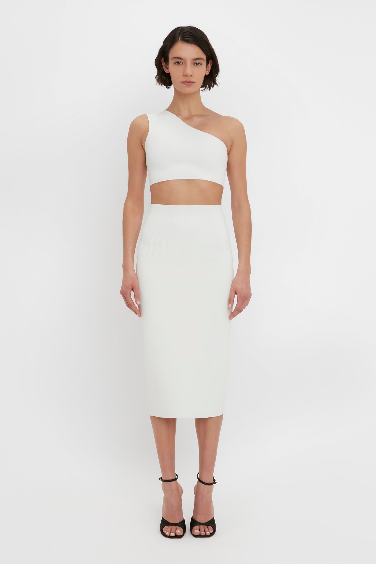 A person stands against a white background wearing a white, one-shoulder, midriff-baring top and the VB Body Fitted Midi Skirt in White by Victoria Beckham, paired with black high-heeled sandals.