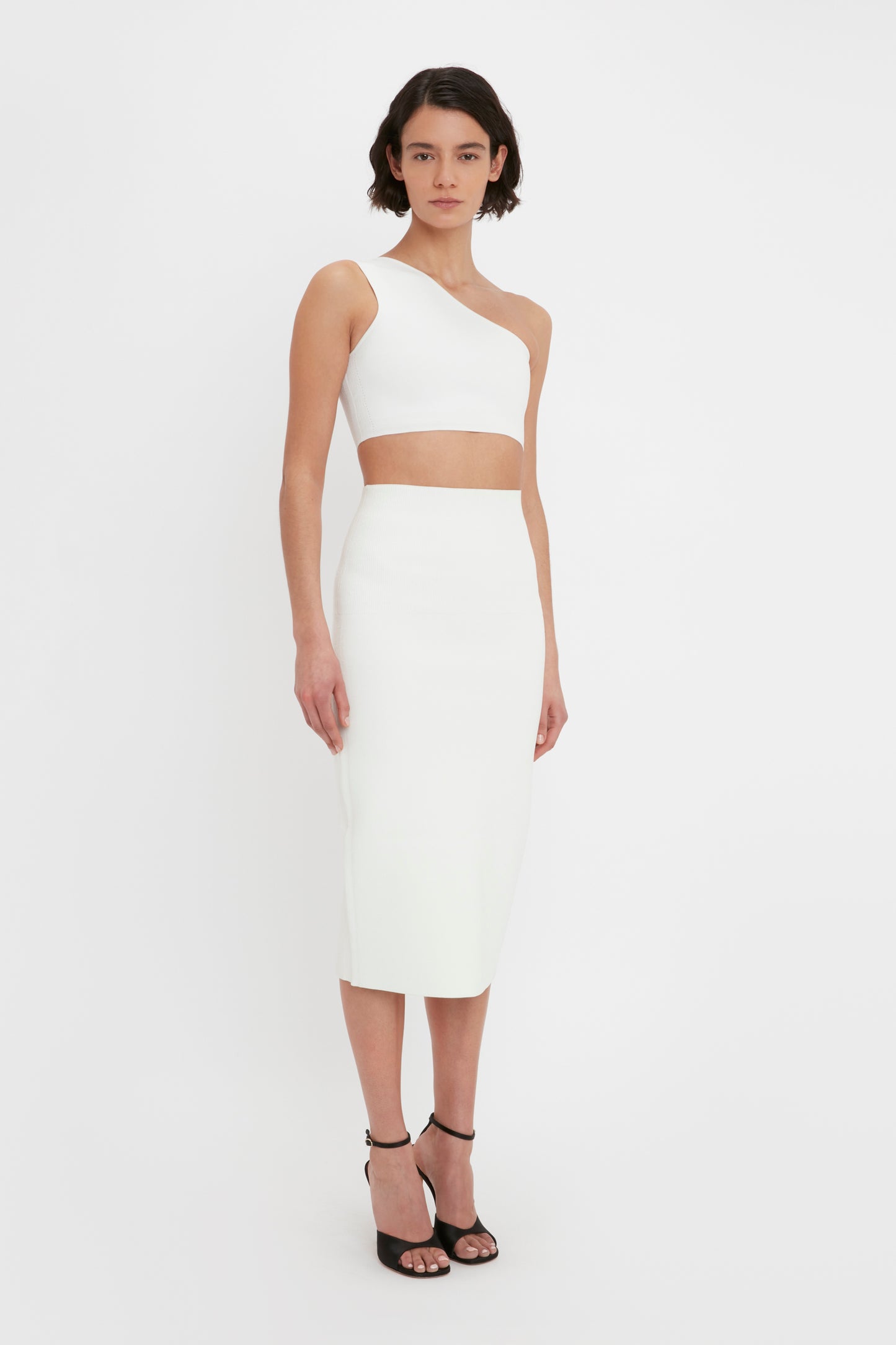 Person wearing a VB Body One Shoulder Crop Top in White by Victoria Beckham and a matching white high-waisted, knee-length skirt, paired with black high-heeled sandals.