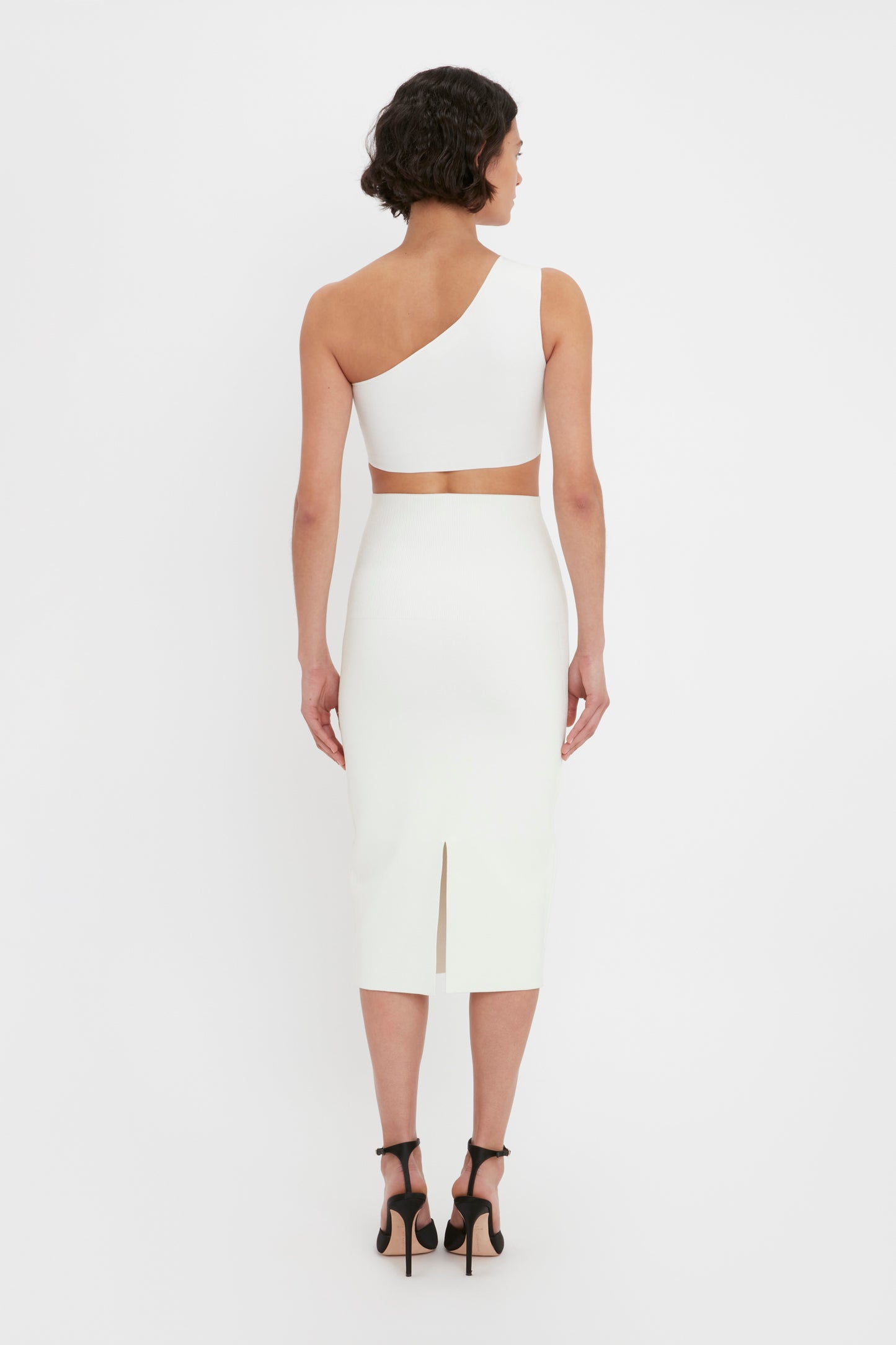 A woman with short dark hair is wearing a Victoria Beckham VB Body One Shoulder Crop Top in white, paired with black high-heeled shoes. She is standing with her back to the camera against a plain white background.
