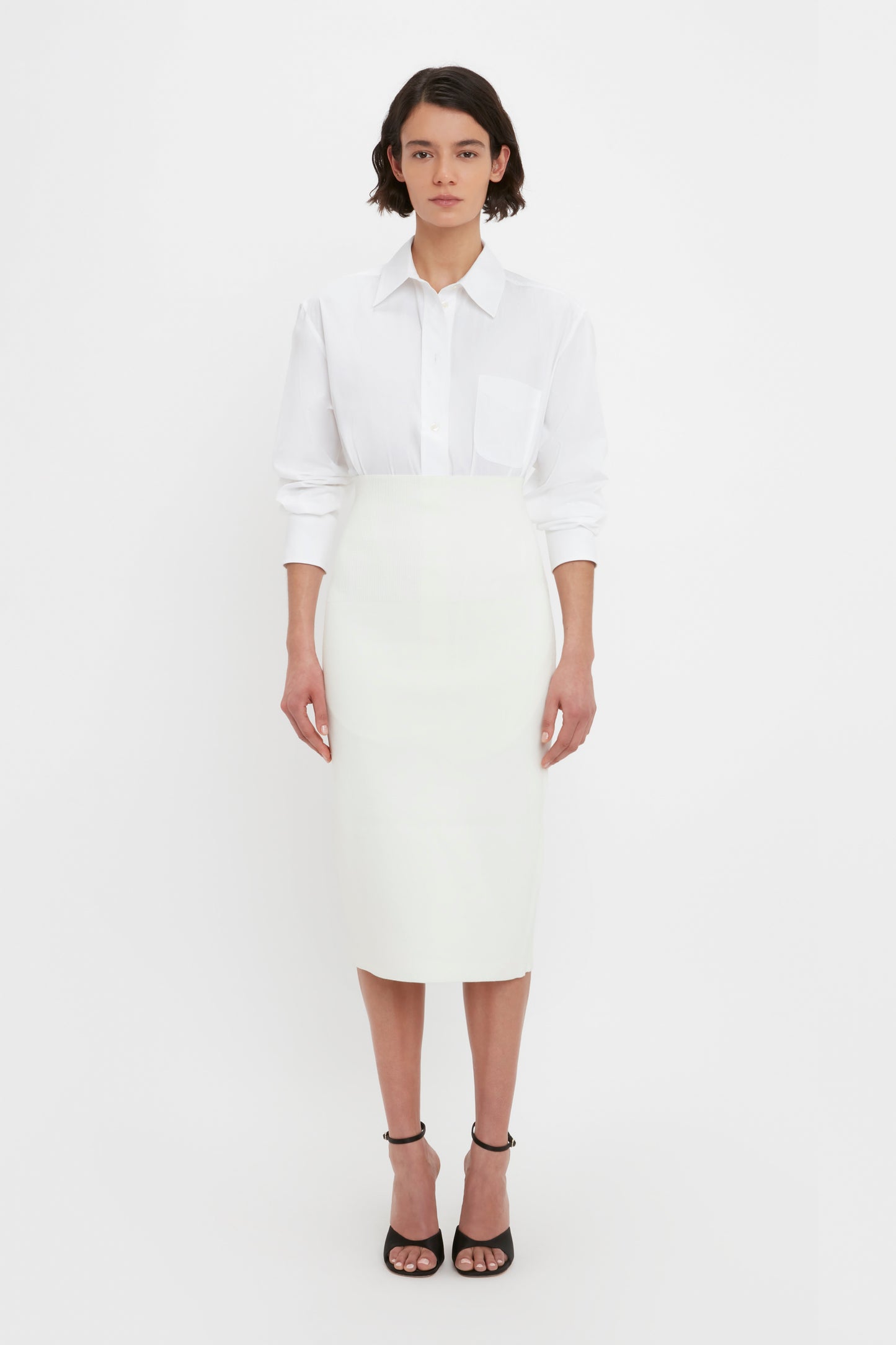 A person is standing against a plain white background, wearing a white long-sleeved shirt, the VB Body Fitted Midi Skirt in White by Victoria Beckham, and black high-heeled sandals.