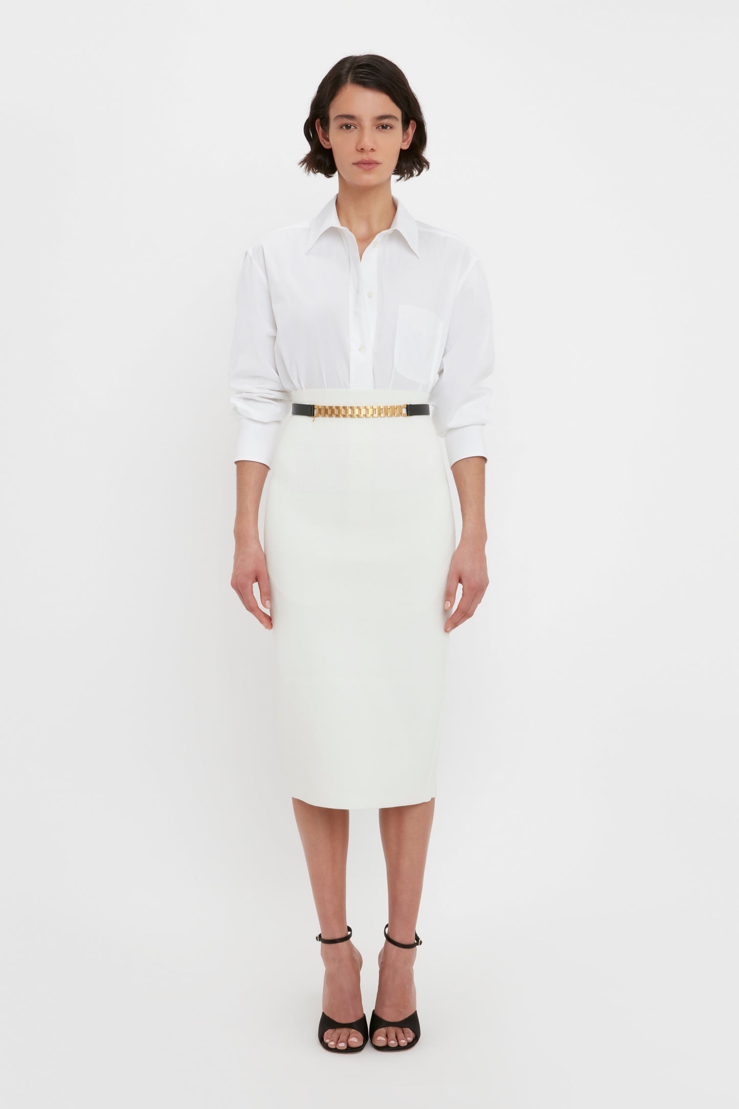 A person stands against a white background wearing a white collared shirt tucked into the "VB Body Fitted Midi Skirt in White" from Victoria Beckham, accessorized with a black belt. They are also wearing black high-heeled shoes.