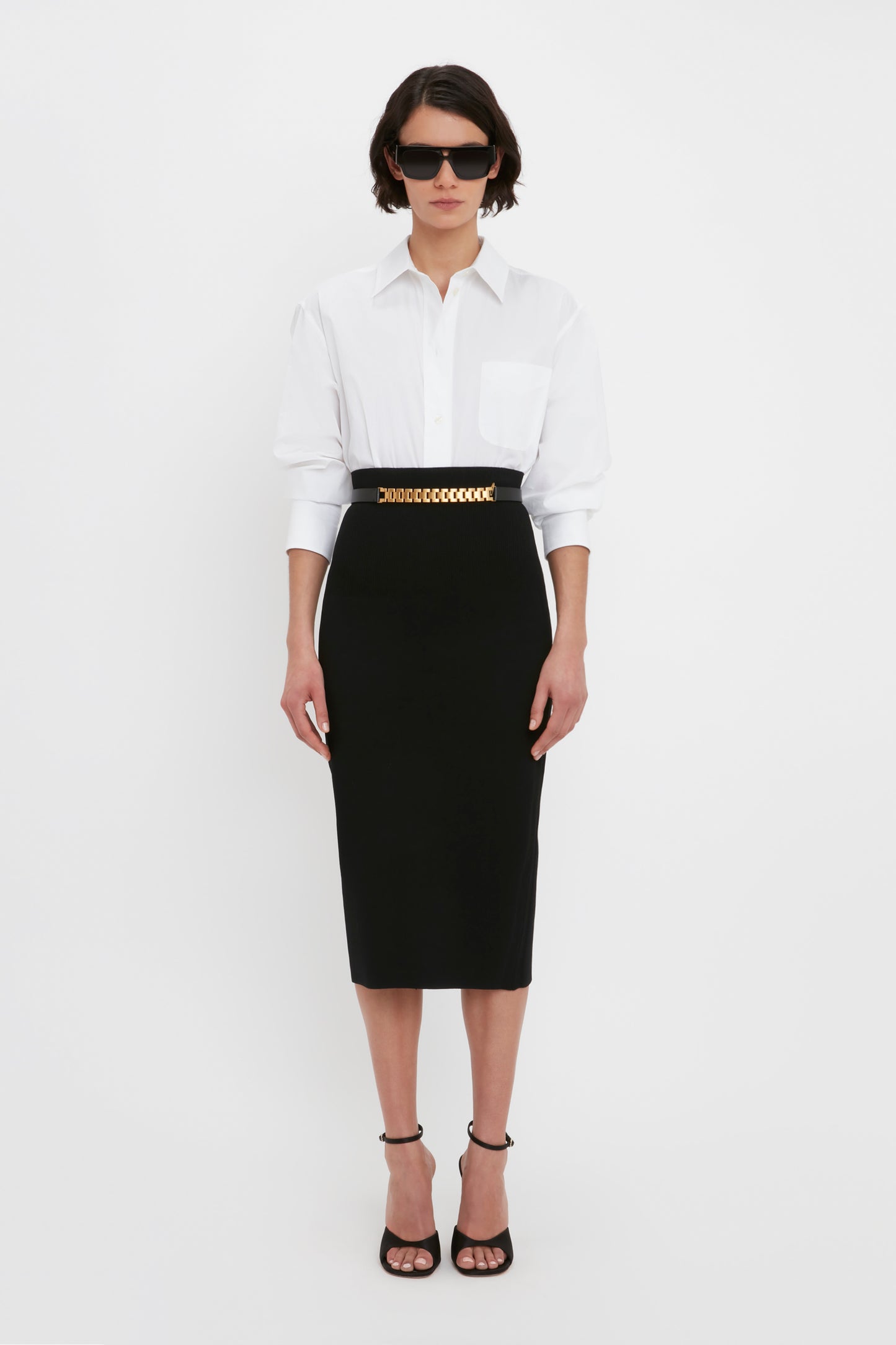 A person stands against a white background wearing sunglasses, a white button-down shirt, a chain belt, black high-heeled sandals, and the VB Body Fitted Midi Skirt in Black from Victoria Beckham.