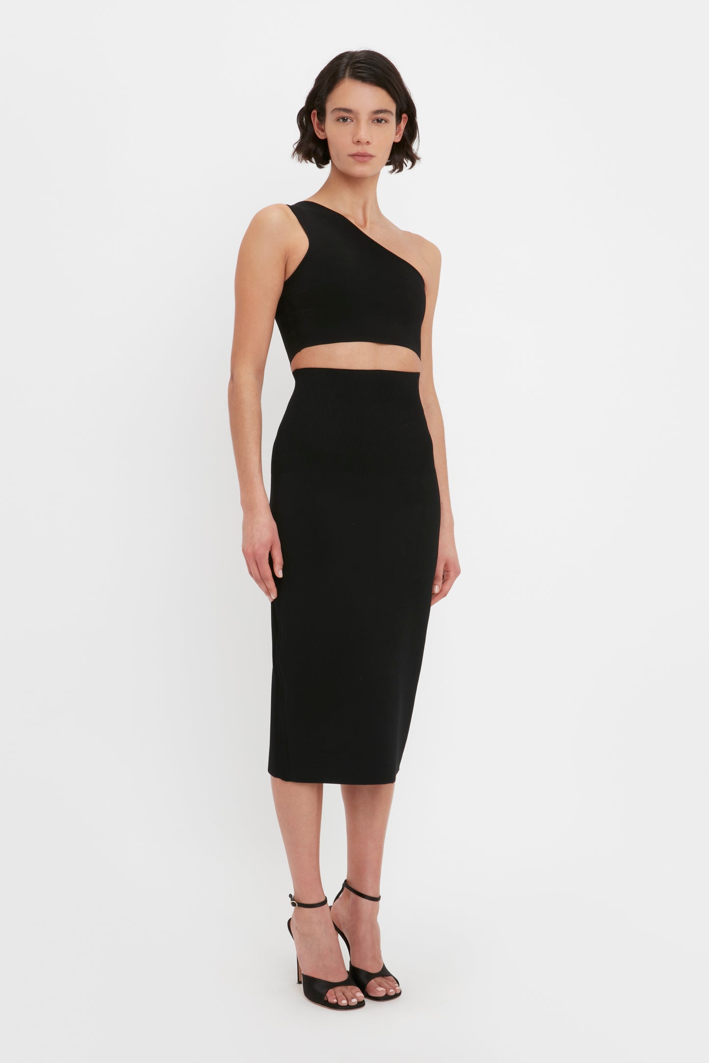 Person standing in front of a plain white background, wearing a black one-shoulder top and the Victoria Beckham VB Body Fitted Midi Skirt in Black, paired with black high-heeled sandals.