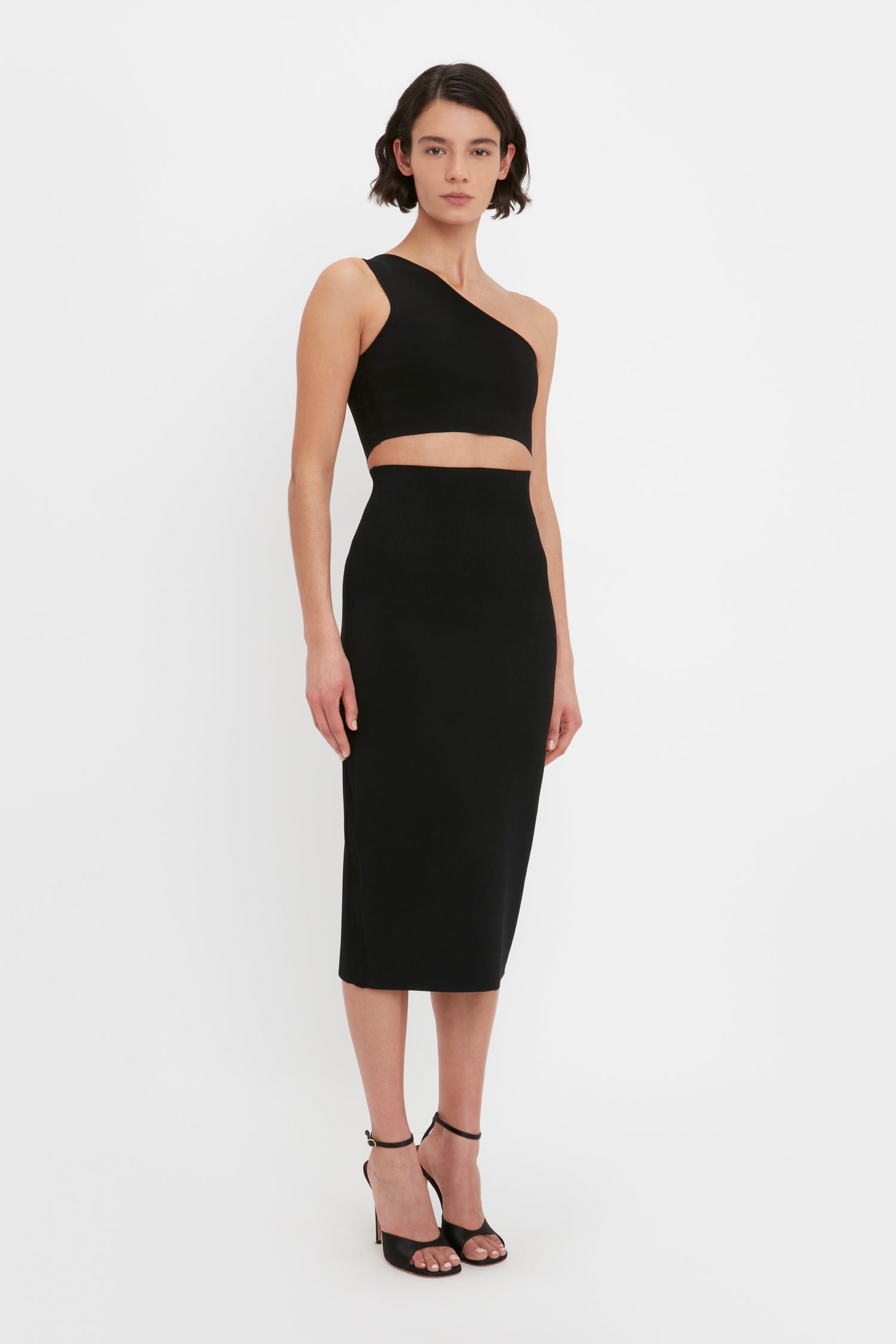 Person standing in front of a plain white background, wearing a black one-shoulder top and the Victoria Beckham VB Body Fitted Midi Skirt in Black, paired with black high-heeled sandals.