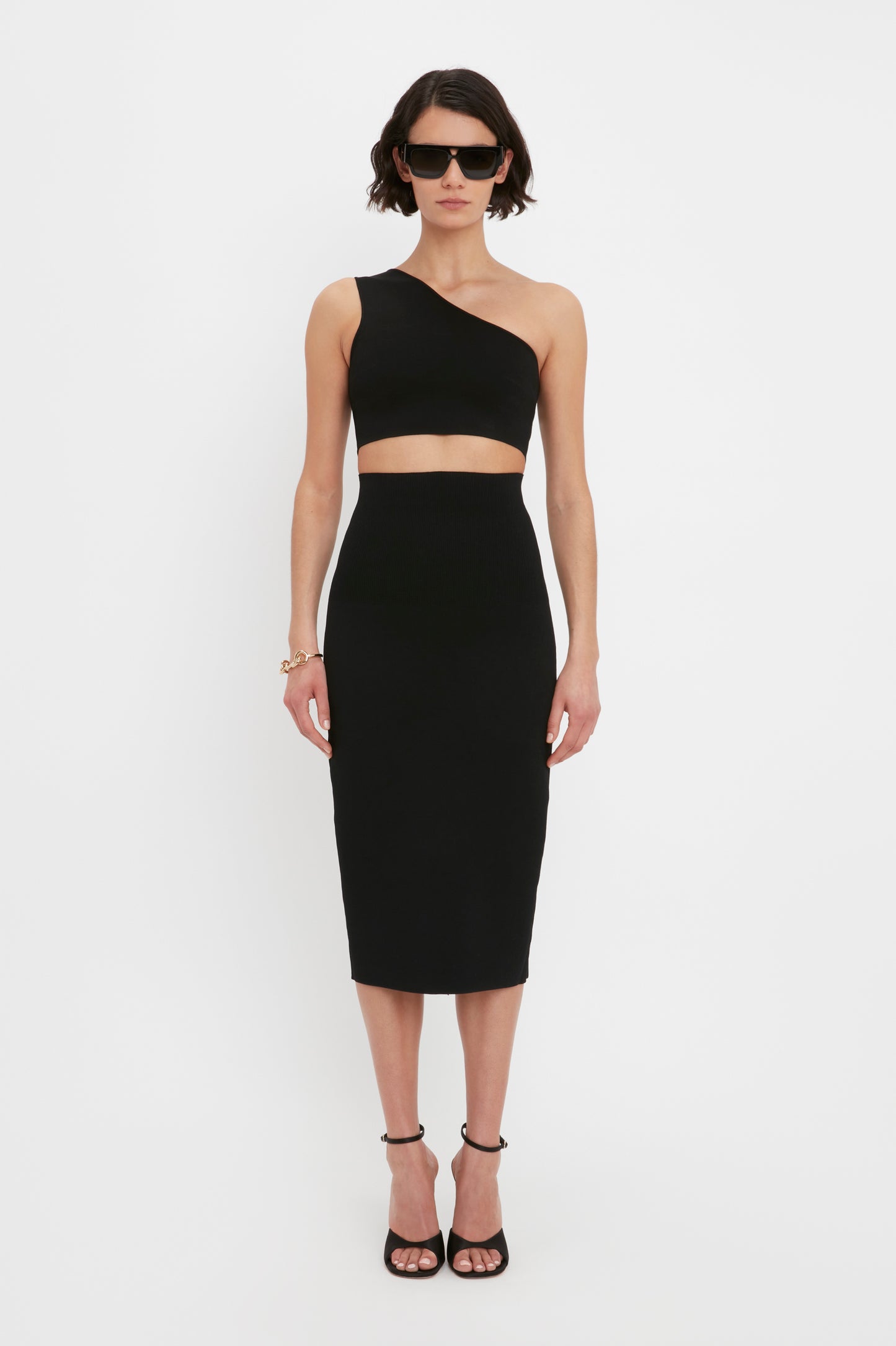 Person standing against a white background wearing a black one-shoulder crop top, the VB Body Fitted Midi Skirt in Black by Victoria Beckham, black high heels, and sunglasses.