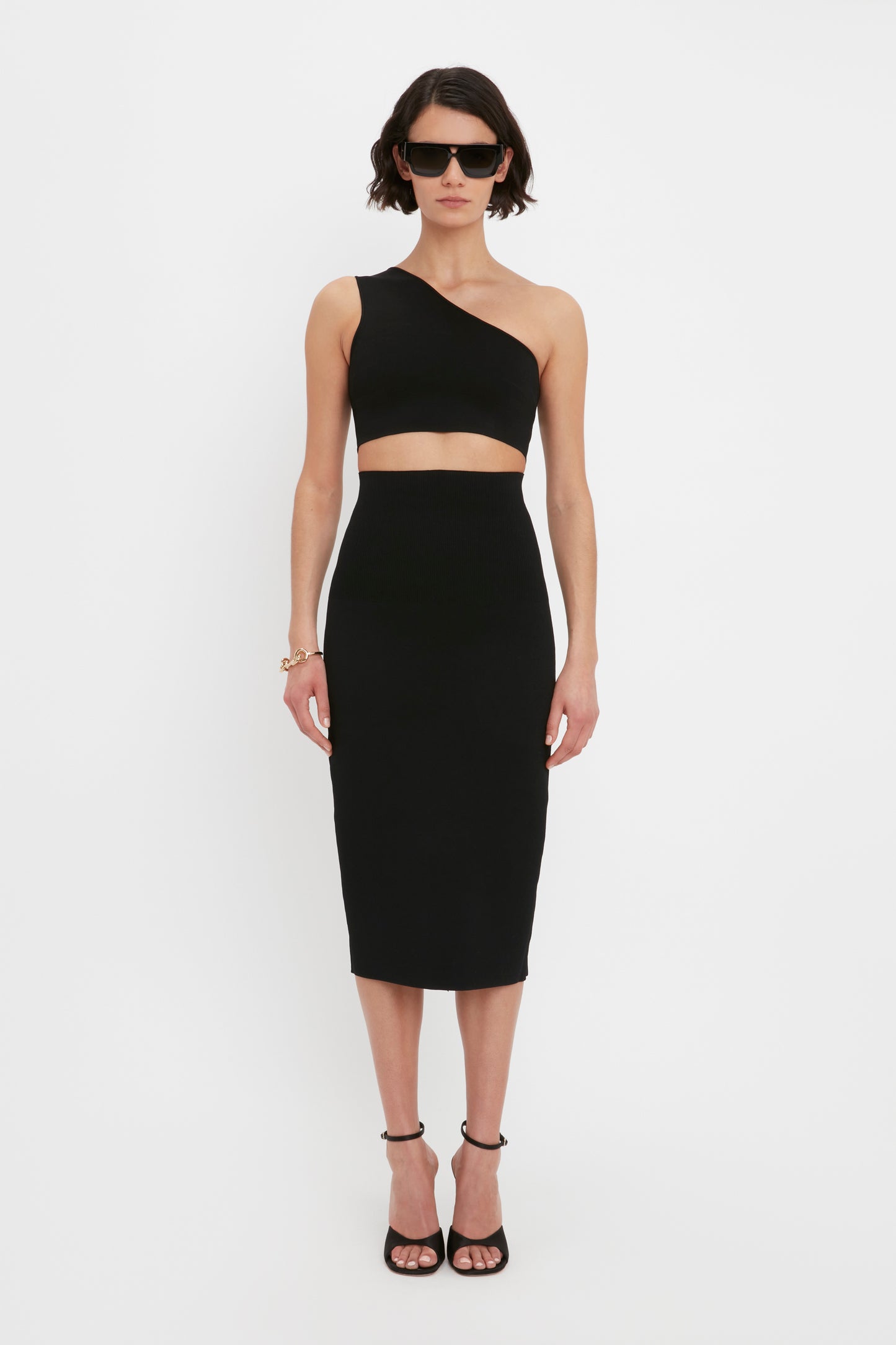 A person wearing sunglasses, the VB Body One Shoulder Crop Top in Black by Victoria Beckham, and a matching high-waisted knee-length skirt with black heels, standing against a plain white background.